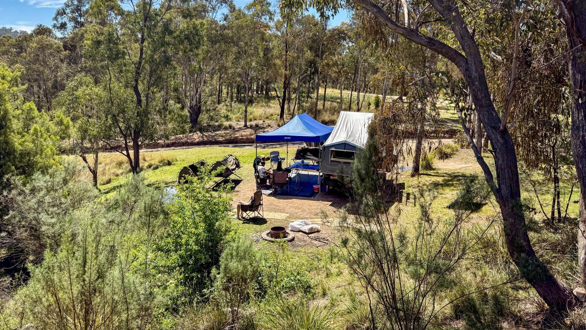 Campers sit under a blue gazebo next to a grey camper trailer surround by the green Australian bush and blue skies above. Image by Melissa Ellison 2025.
