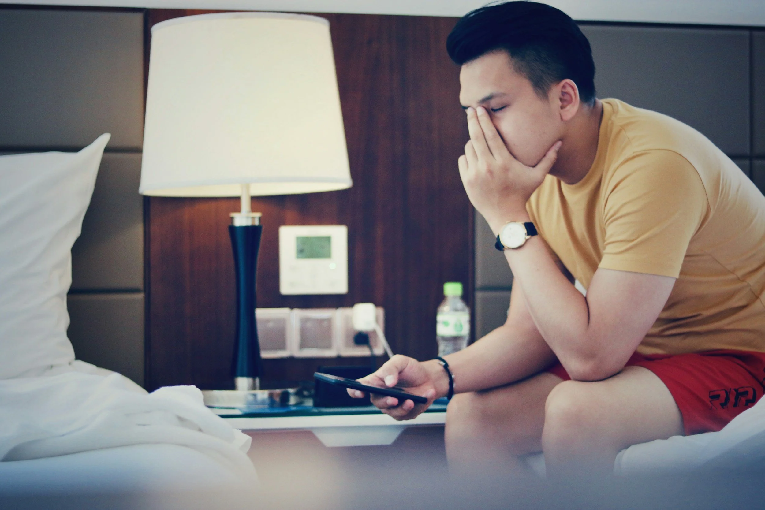 A young gay man looking worried, phone in hand and head in hand, illustrating the anxiety and self-monitoring that comes with approval seeking in relationships