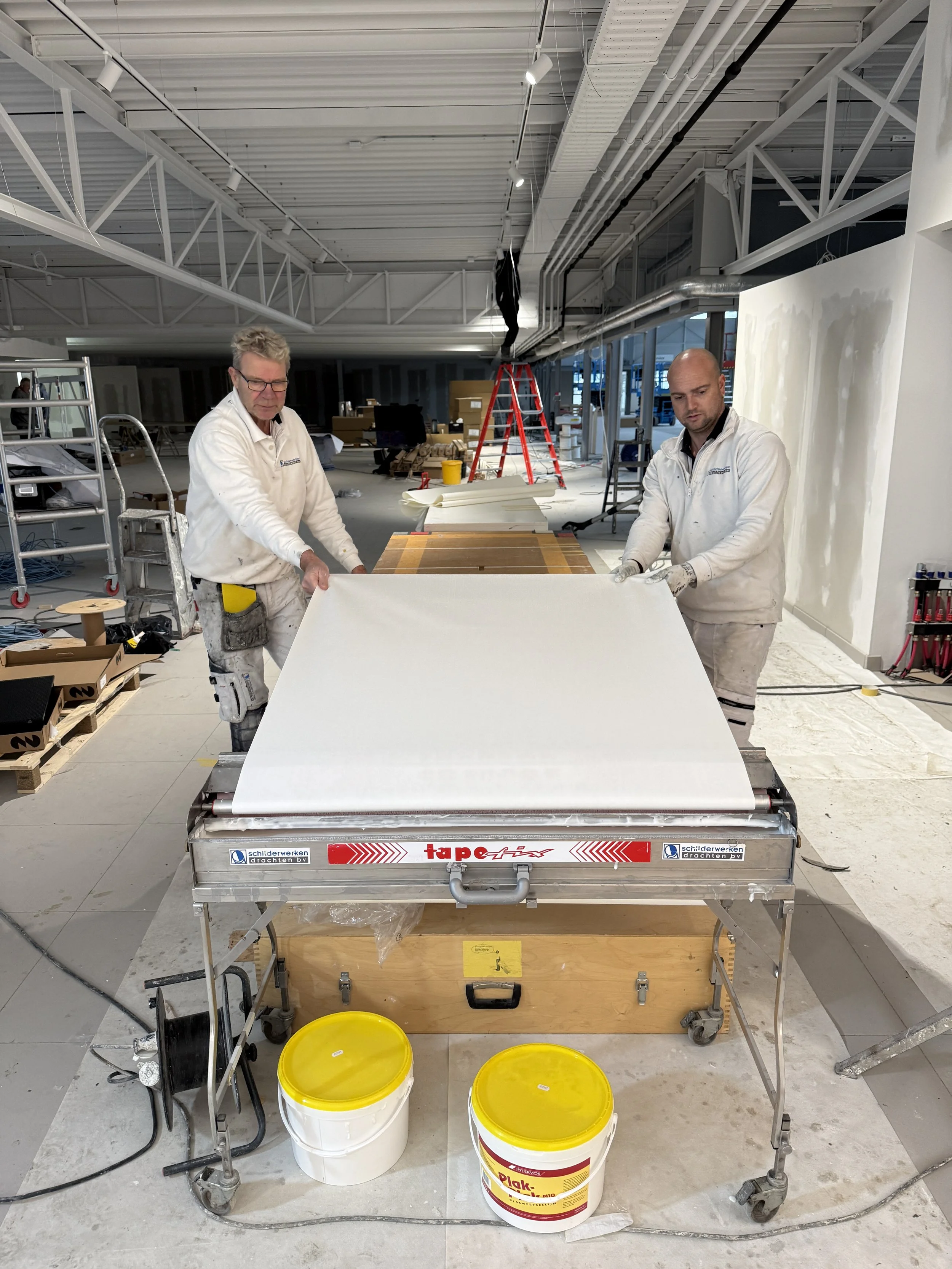 Twee bouwvakkers werken in een interieur, ze bewegen een groot wit paneel op een metalen tafel, op de achtergrond bevinden zich ladders, gereedschap en bouwmaterialen.