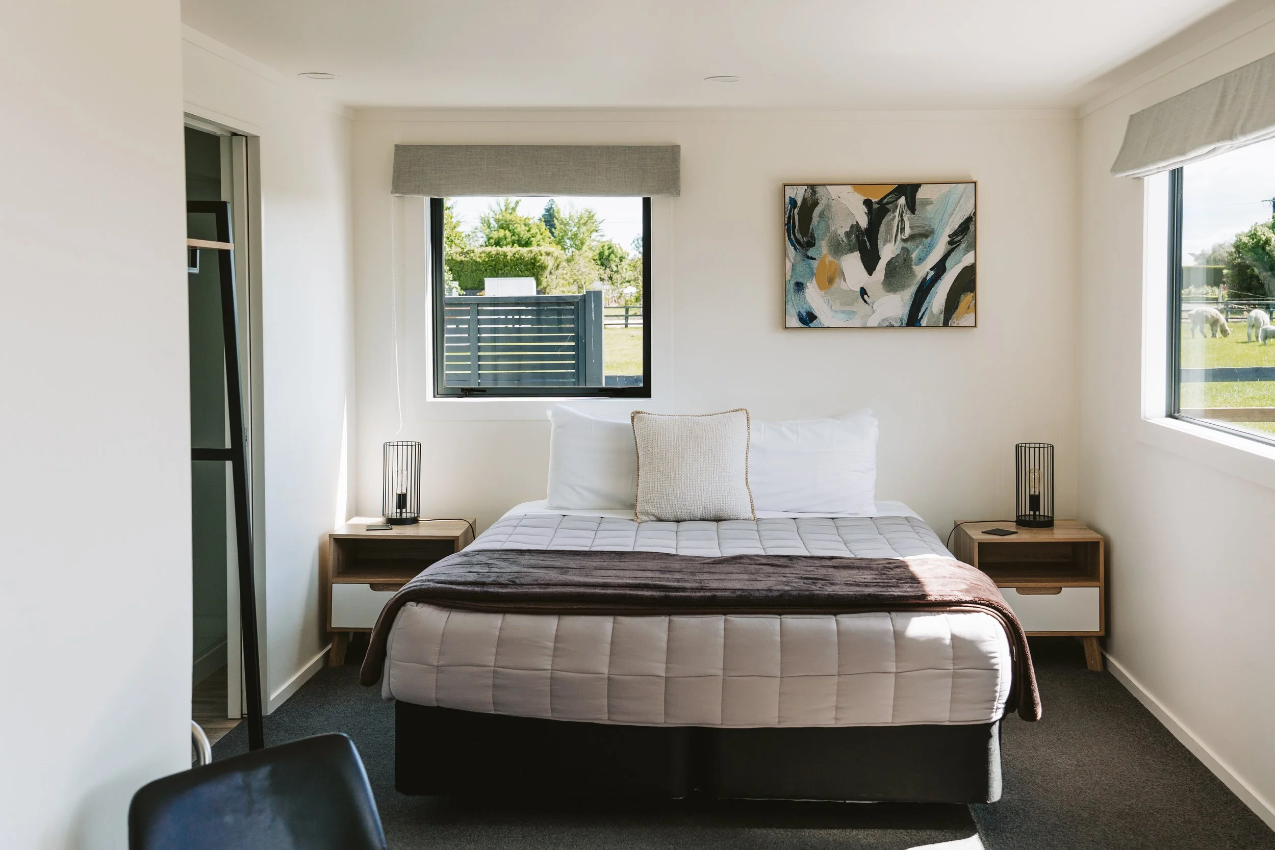 Bedroom with a bed, two side tables with lamps, abstract artwork on the wall, two windows showing outdoor greenery.