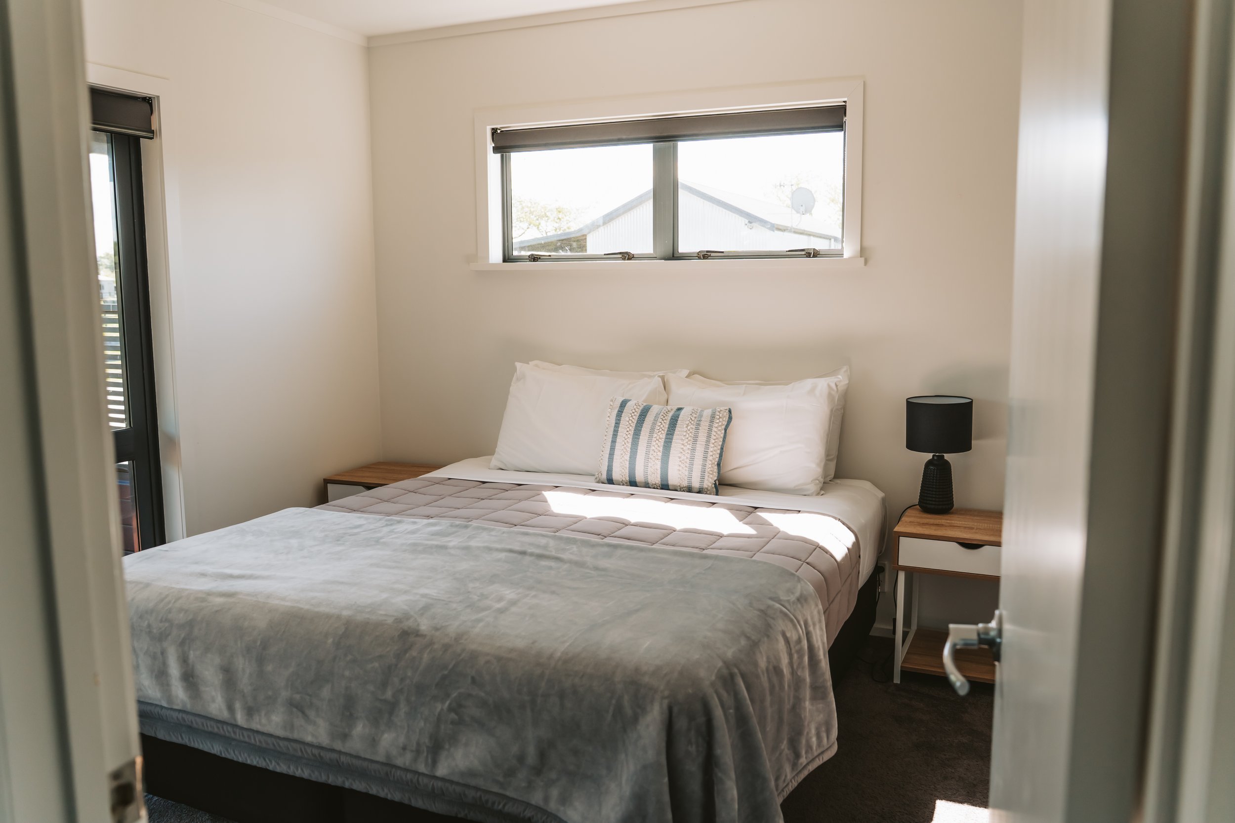 Bright and inviting bedroom. Featuring a comfortable King bed with crisp white linen, soft grey bedding, bedside tables with modern lamps, and large windows allowing natural light to fill the room.