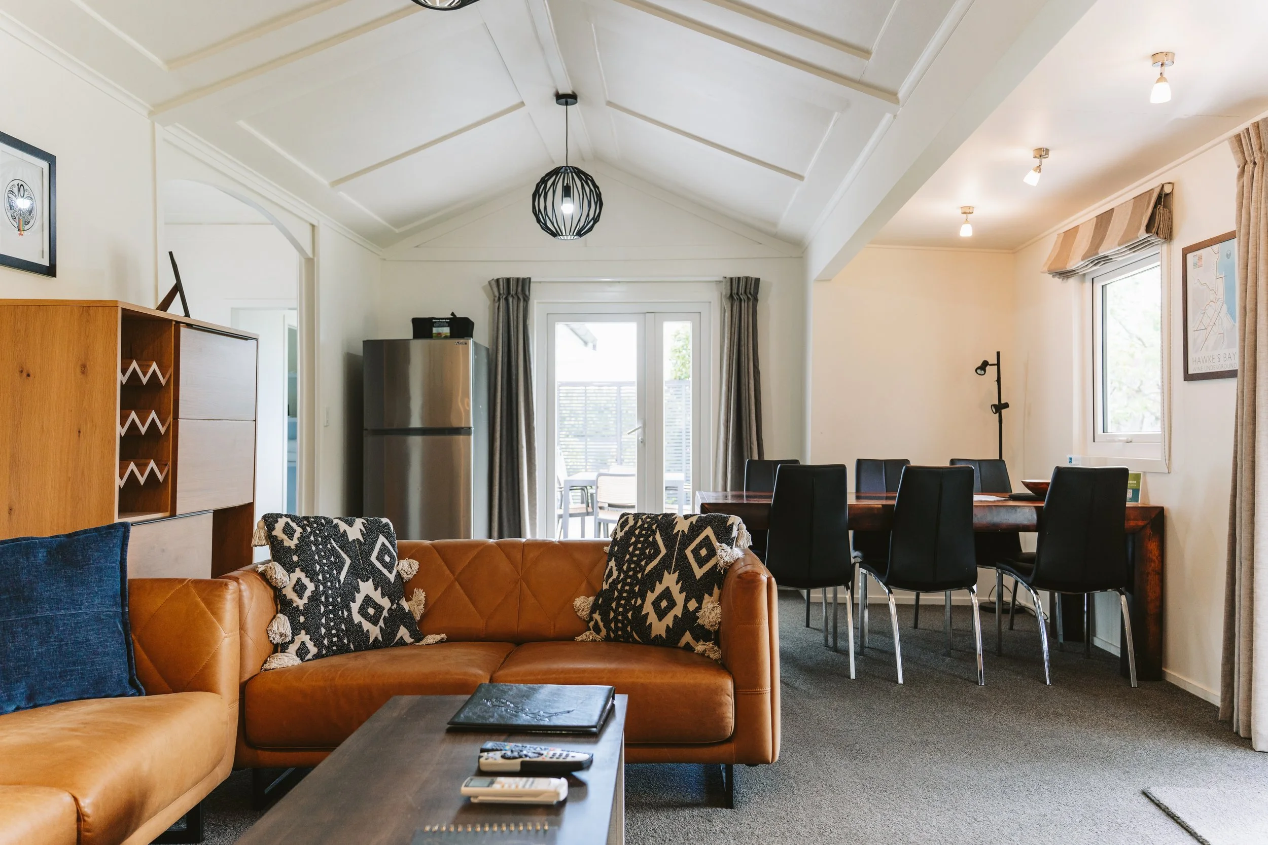 Spacious open-plan living and dining area inside a modern cottage. Features include leather sofas with patterned cushions, a large wooden dining table with seating for eight, vaulted ceilings with modern lighting, soft neutral décor, and French doors