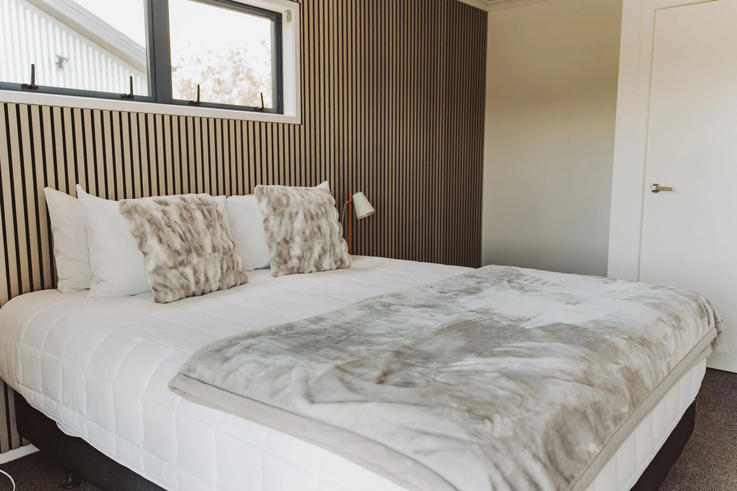 A neatly made bed with a white quilt, two white pillows, and two decorative beige pillows, with a green throw blanket at the foot of the bed, in a minimalist bedroom with white walls, black curtains, a window, two white nightstands with lamps, a wall mirror, and a sliding glass door leading to a balcony.