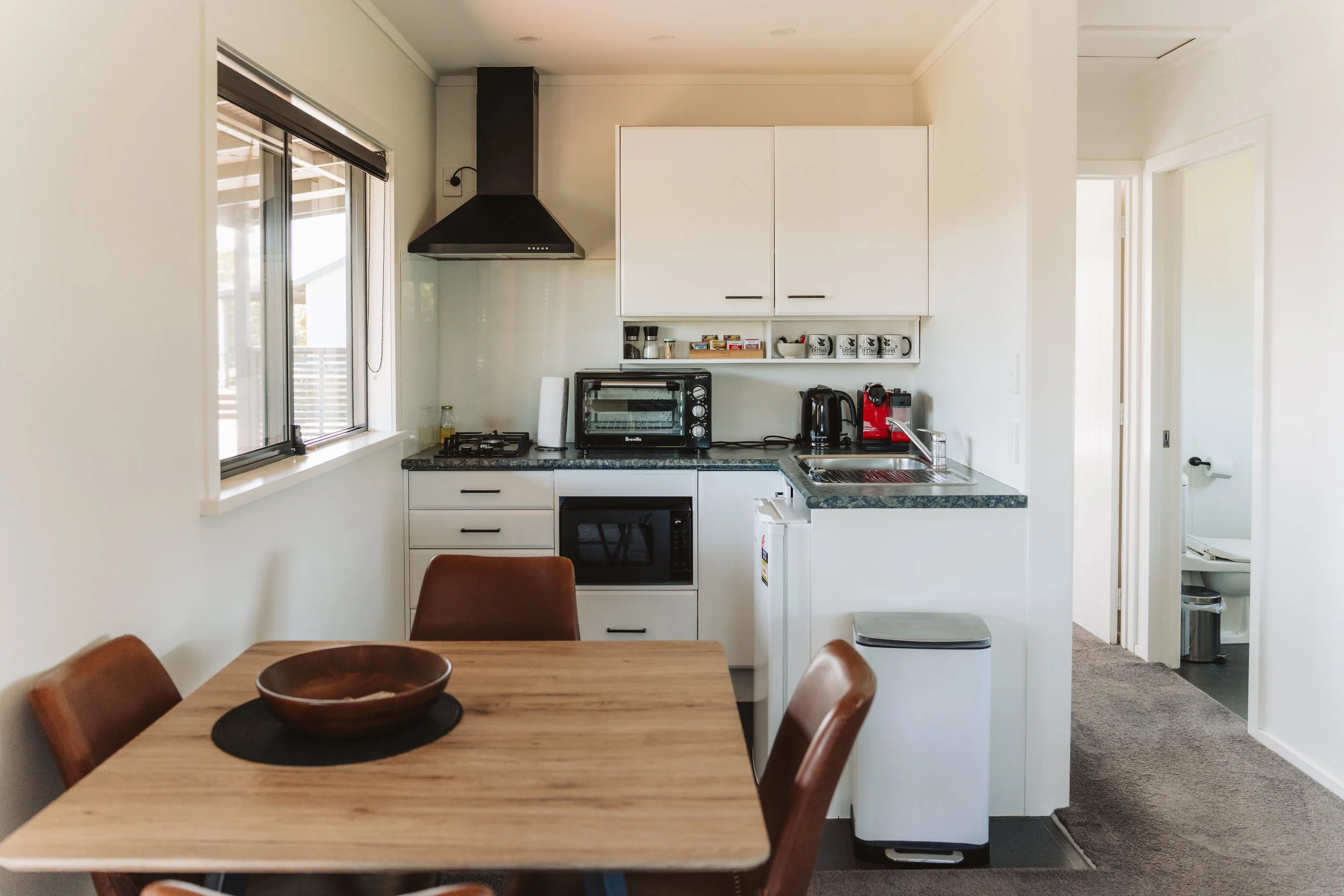 Modern and light-filled kitchenette and dining area, Features include a compact fully equipped kitchen with cooktop, oven, microwave, coffee machine, kettle, dining table with leather chairs, natural light through large windows.