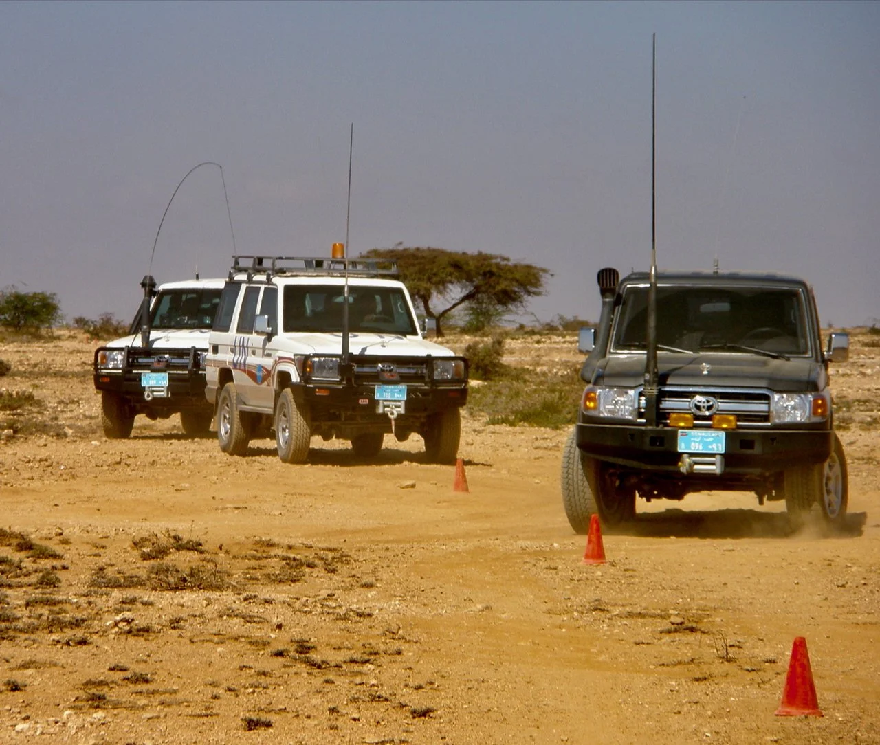Three off-road vehicles driving on a dirt trail in a desert landscape, with orange cones marking the course and a tree in the background under a cloudy sky.