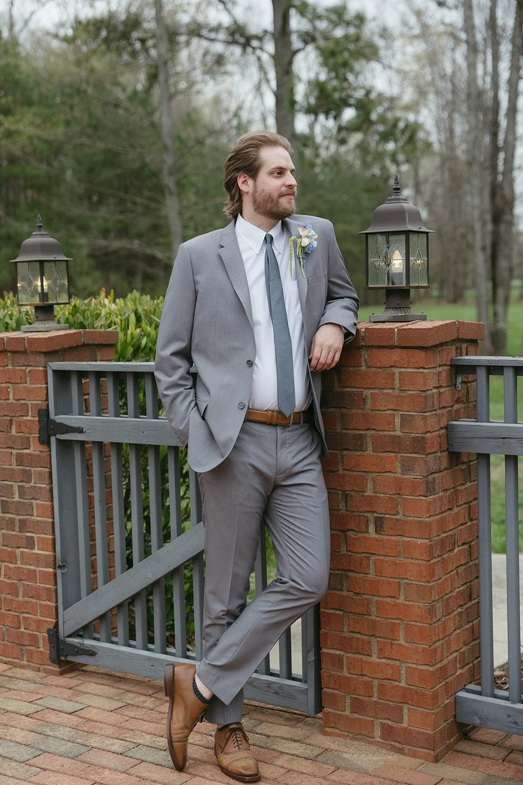 A man in a gray suit with a blue tie and a boutonniere leans against a brick pillar with lanterns, outdoors with trees and greenery in the background.