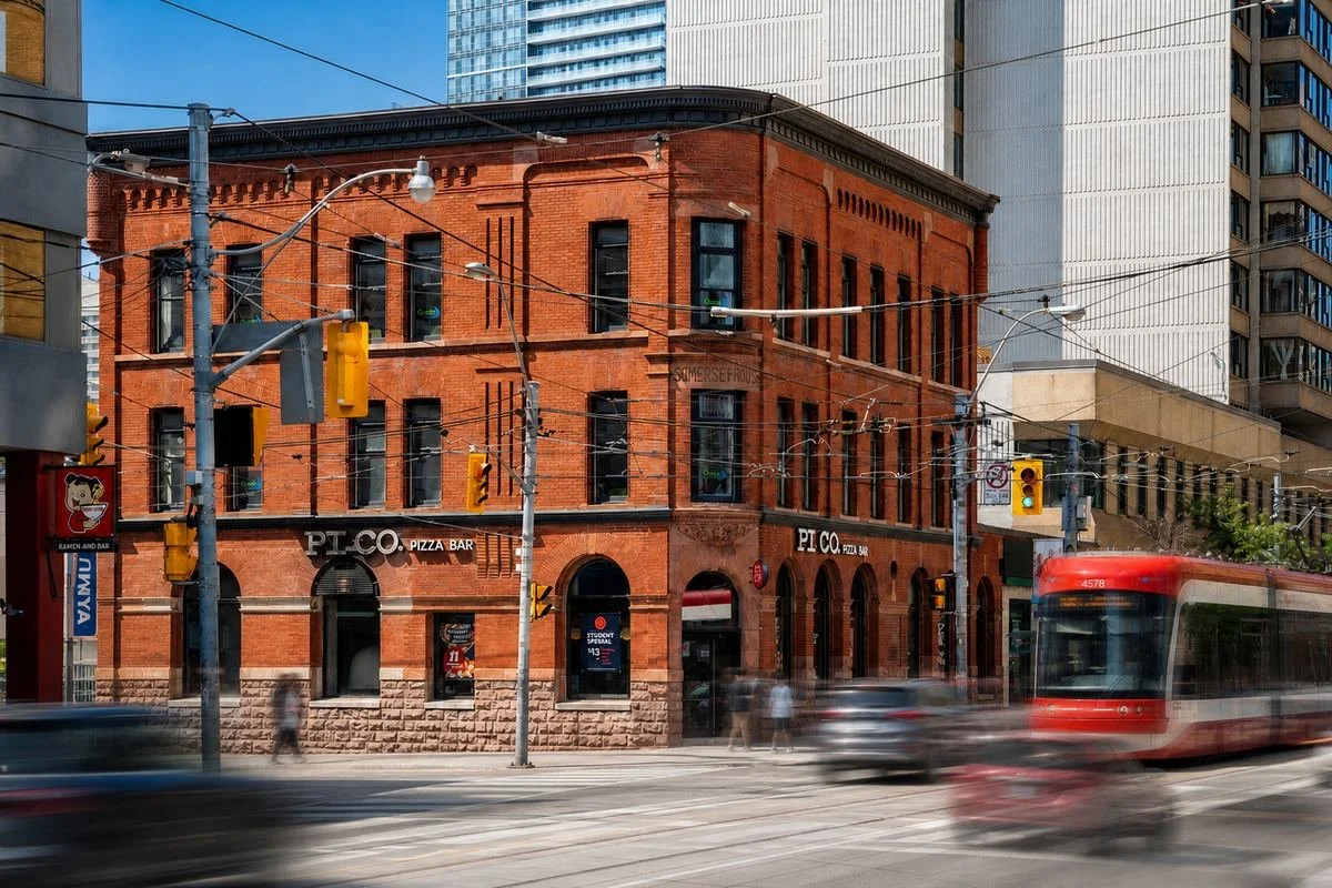 A busy city street with a red brick building housing pizza and bar establishments, a streetcar moving quickly, traffic lights, and pedestrians crossing the street in daytime.