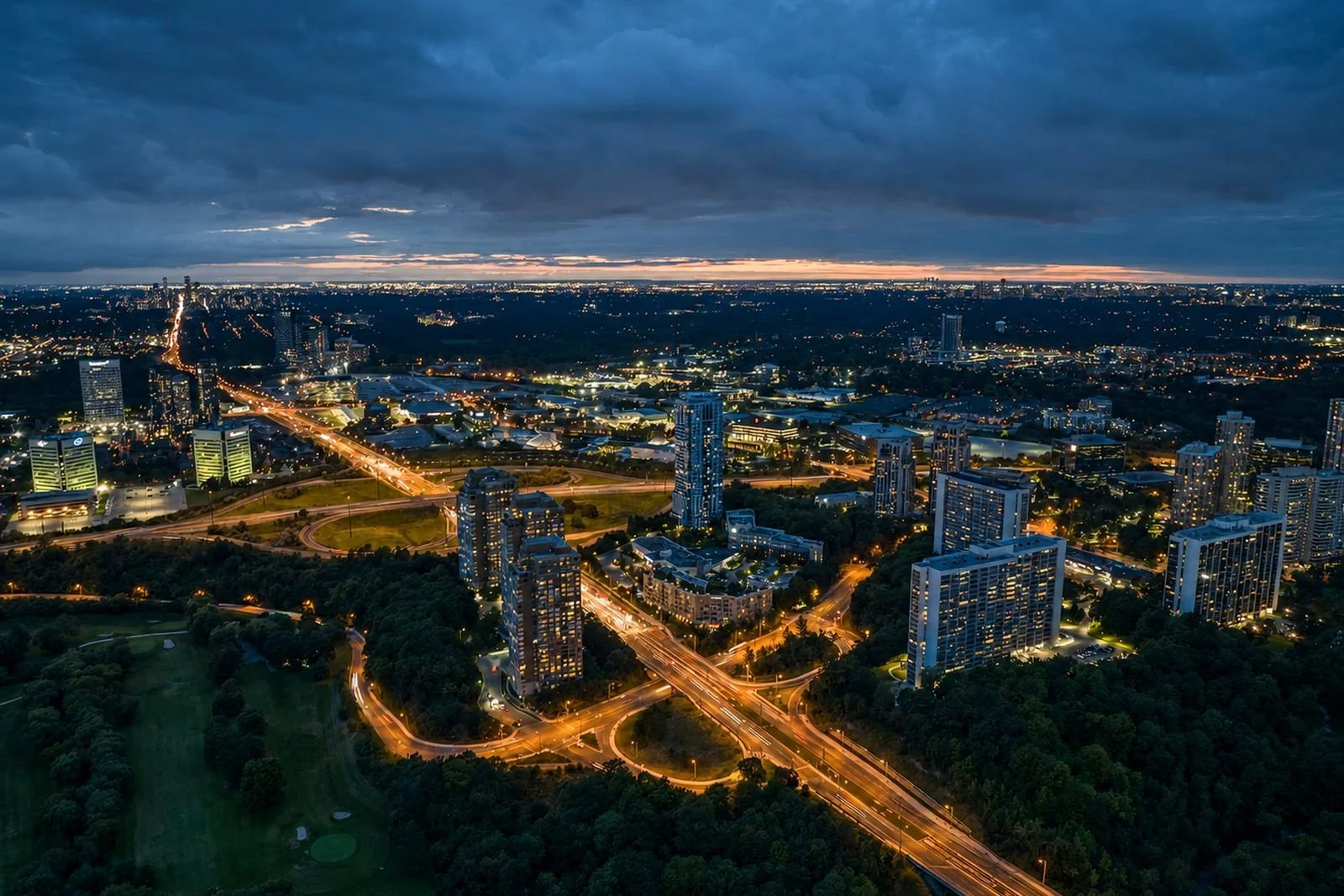 An aerial view of a city at dusk with illuminated roads and skyscrapers, and a cloudy sky.
