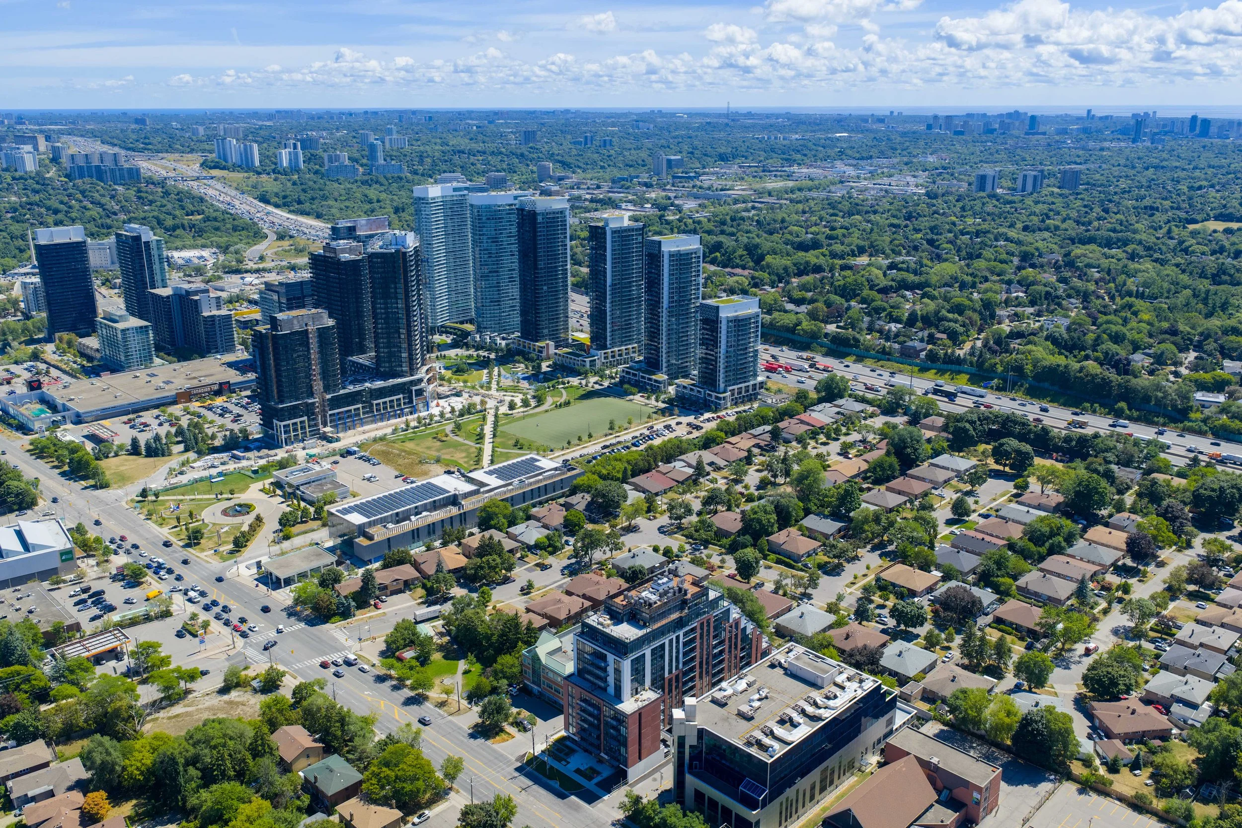 Aerial view of a cityscape showing a cluster of modern high-rise buildings alongside residential houses, with green spaces and trees interspersed, under a partly cloudy sky.