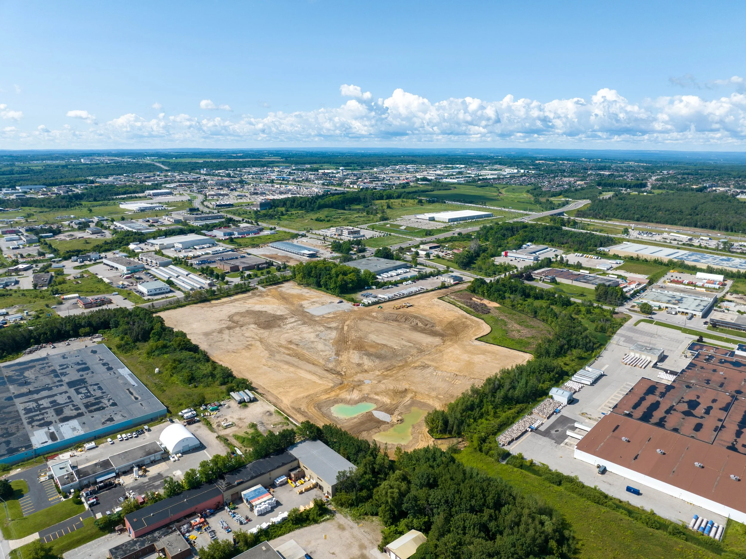 An aerial view of an industrial park with warehouses, commercial buildings, roads, and some areas of cleared land and greenery under a blue sky with scattered clouds.