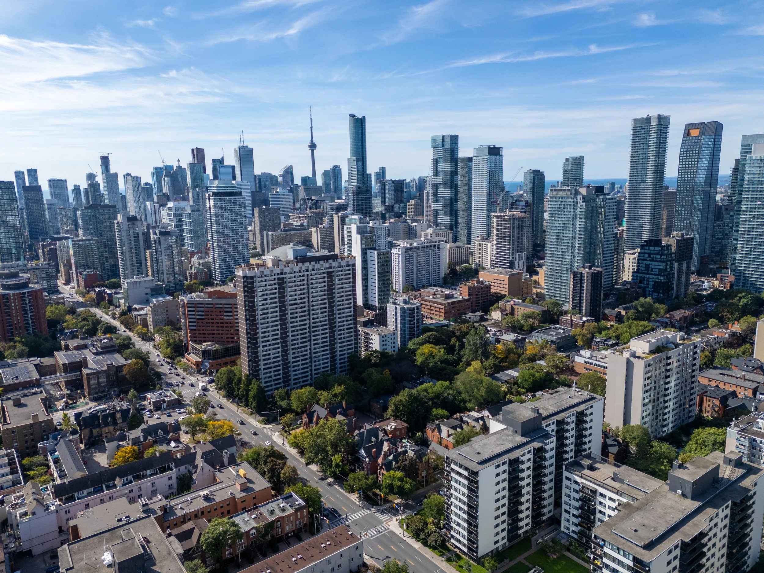 A cityscape featuring tall skyscrapers and buildings under a partly cloudy sky, with a mix of modern and older architecture, and streets lined with trees and smaller structures.