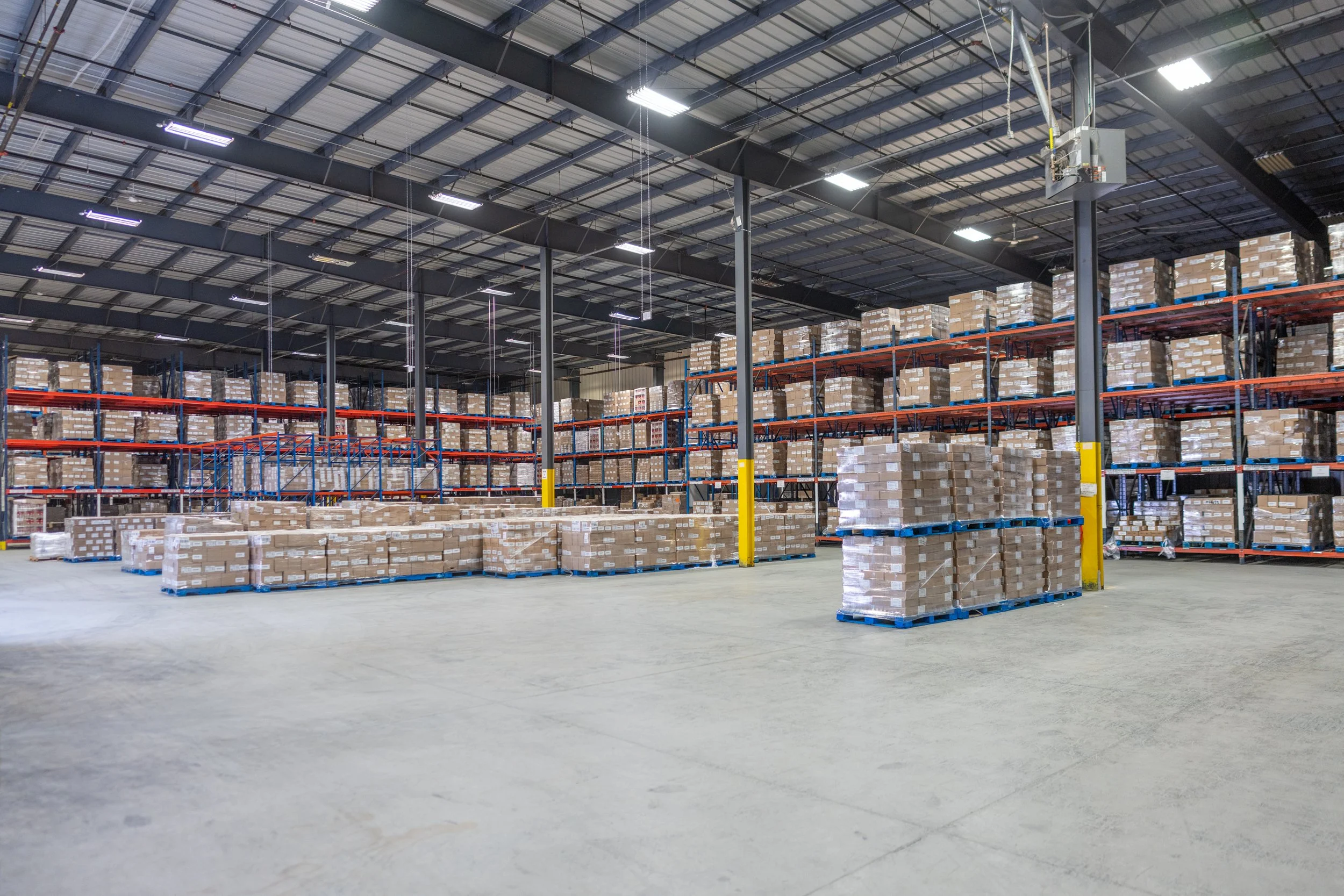 Interior of a large warehouse with industrial shelving filled with cardboard boxes, concrete floor, and a ceiling with metal beams and fluorescent lights.