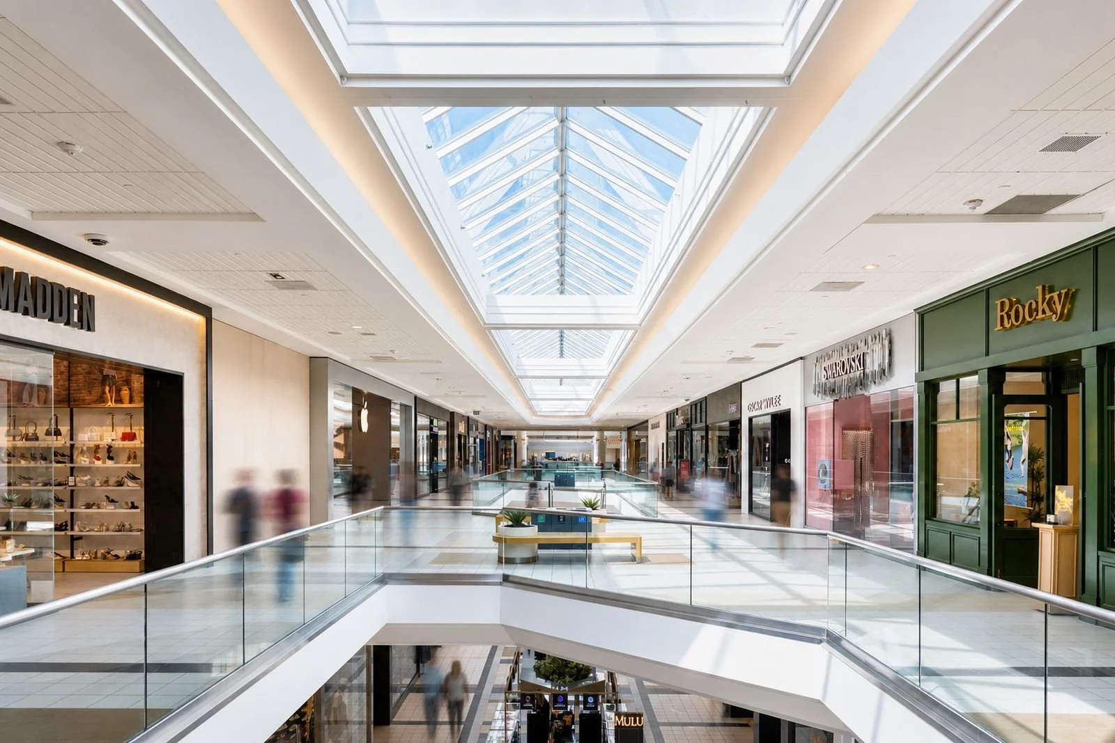 Interior of a modern shopping mall with multiple stores, glass ceiling skylights, and people walking.