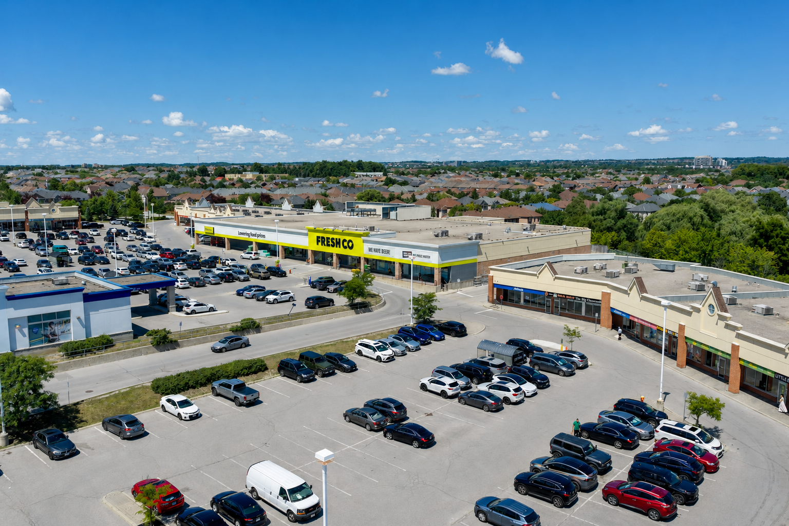 An aerial view of a shopping center parking lot with many cars and stores, including a FreshCo supermarket.