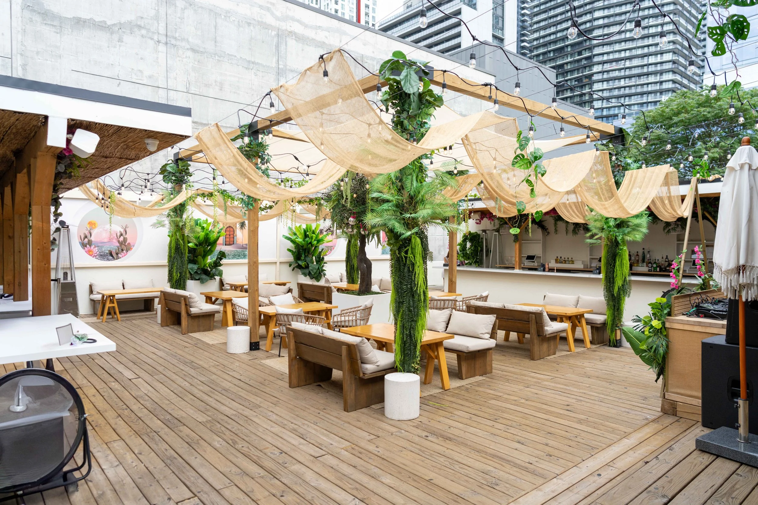 An outdoor patio with wooden flooring, decorated with hanging fabric drapes, string lights, and numerous green plants. There are wooden and cushioned seating arrangements, including benches and chairs, and a bar area in the background. Tall buildings