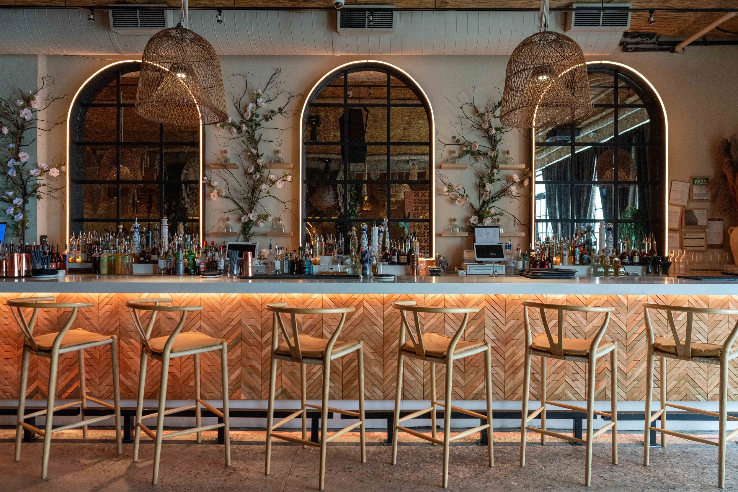 Modern bar with a white countertop, wooden barstools, three arched mirrors with plant and flower decorations, wicker lamps, and a back wall with shelves of bottles, illuminated with warm lighting.