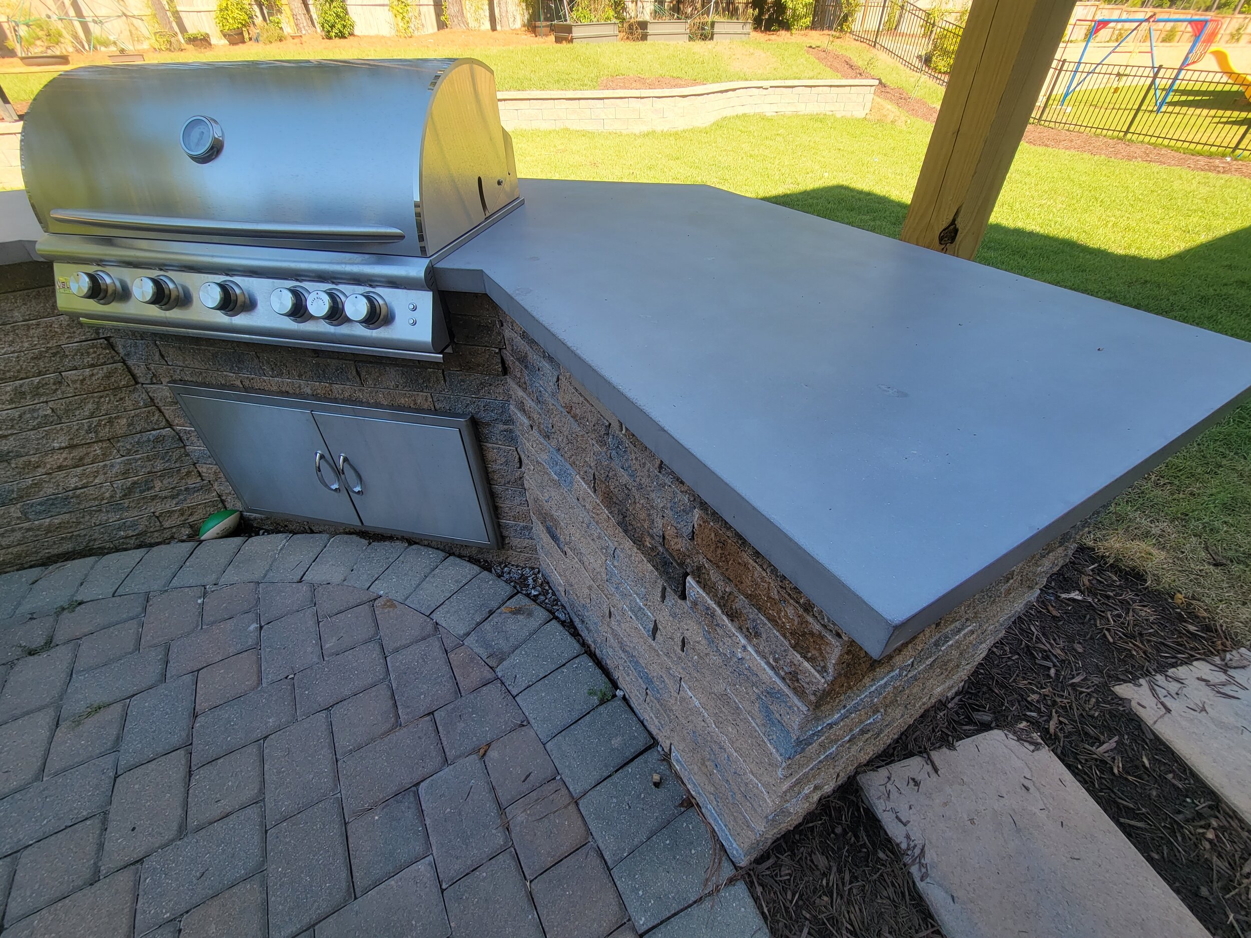 Outdoor kitchen with a stainless steel grill, a concrete countertop, and a brick base, set on a paved patio in a backyard.