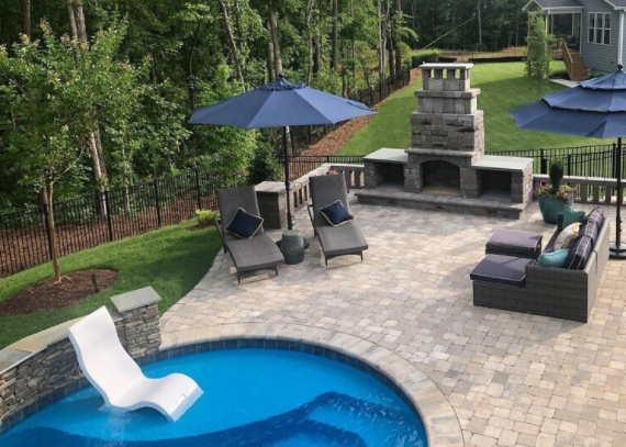 Backyard patio with a swimming pool, lounge chairs with umbrellas, a stone fireplace, and outdoor furniture.
