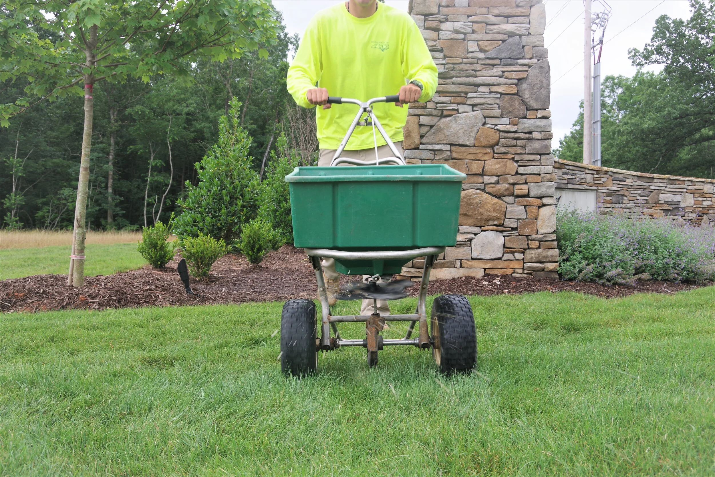 Person pushing a green garden cart on a well-maintained lawn, with bushes, trees, and a stone wall in the background.