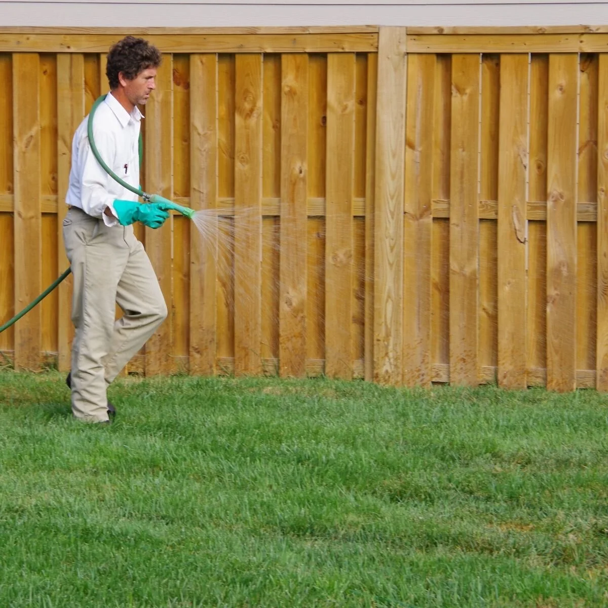 A man in a white shirt, beige pants, and green gloves is spraying liquid on a wooden fence in a backyard with green grass.