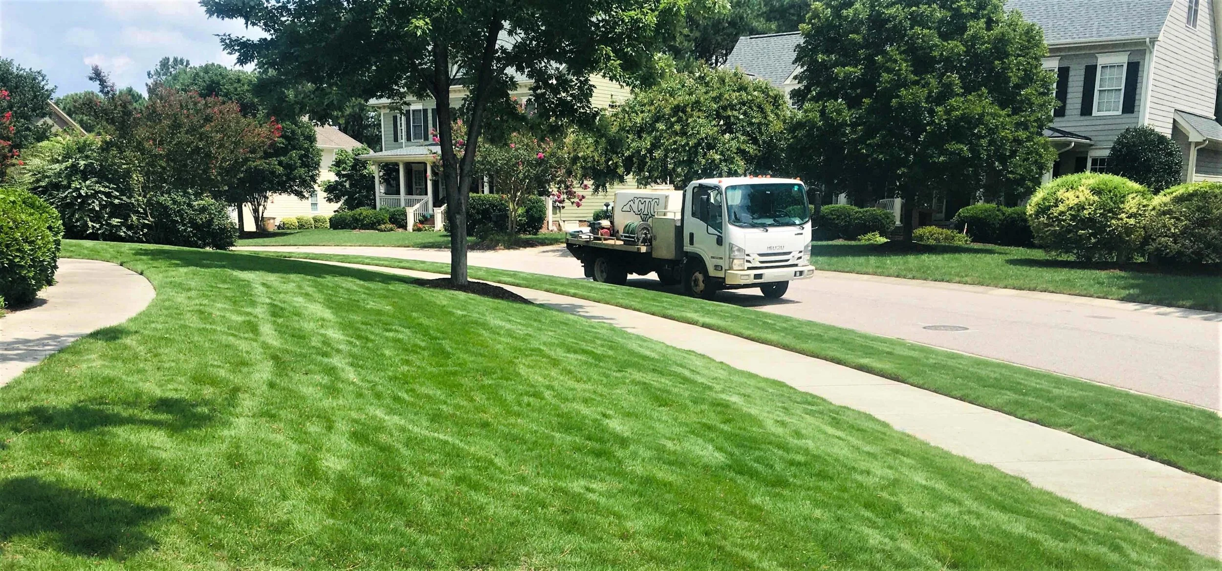 A residential neighborhood with a well-maintained grassy lawn, paved walkway, trees, and houses. A small white service truck is parked on the street.