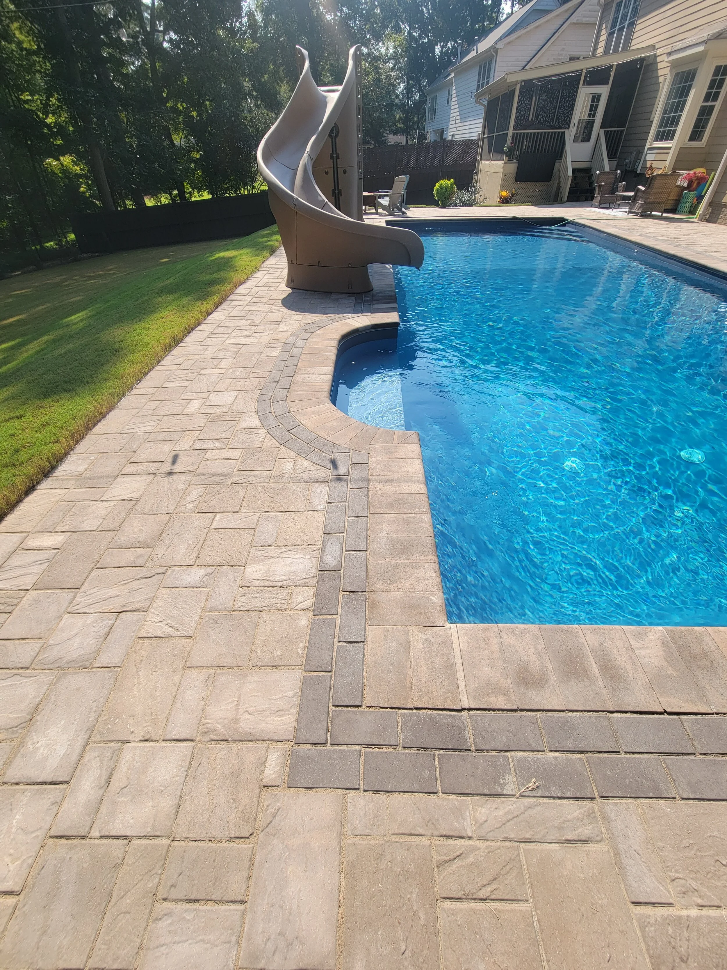 Backyard with a swimming pool and a plastic water slide, surrounded by a stone-paved patio and lawn, with a house and patio furniture in the background.