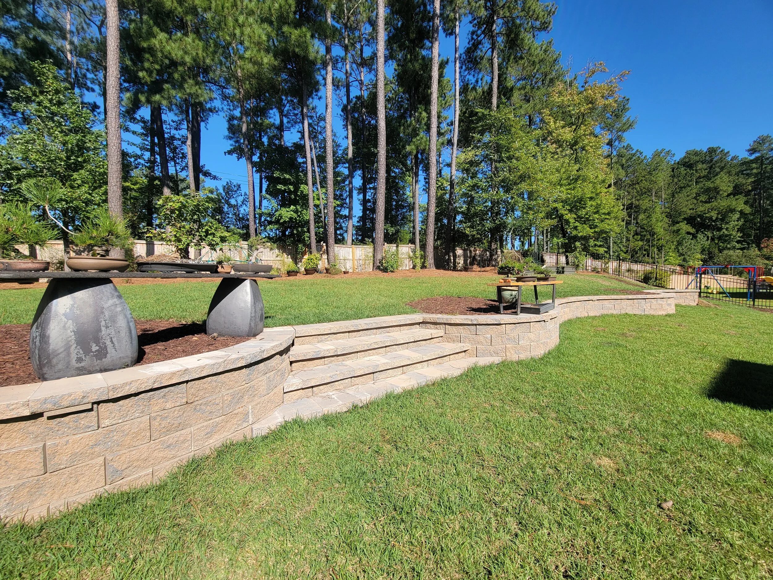 Backyard with tiered stone retaining wall, steps, plates, and a grassy yard with trees and a playground in the background.