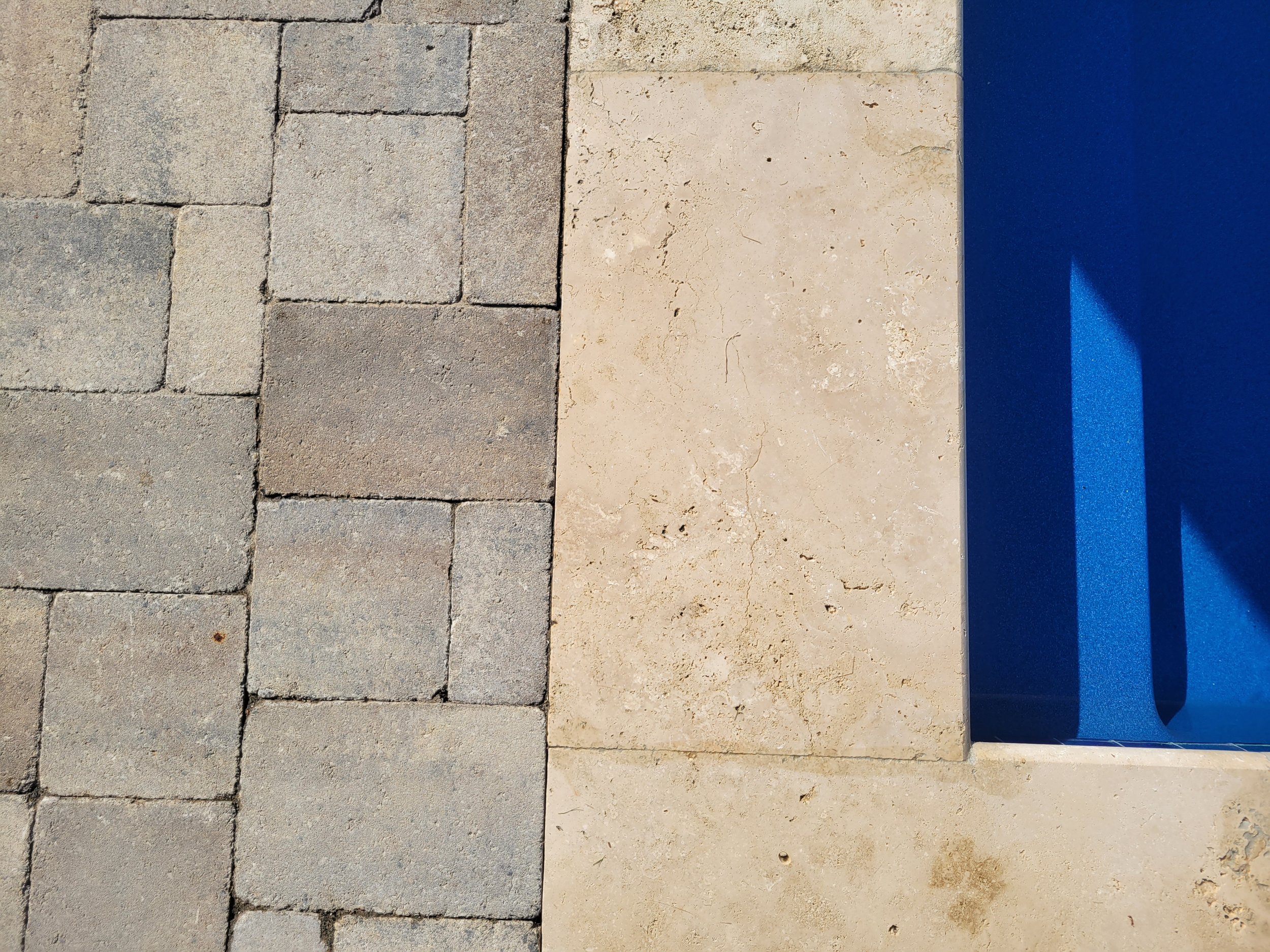 Close-up of a paved stone pathway, a light-colored stone wall, and a section of a blue object casting a shadow.