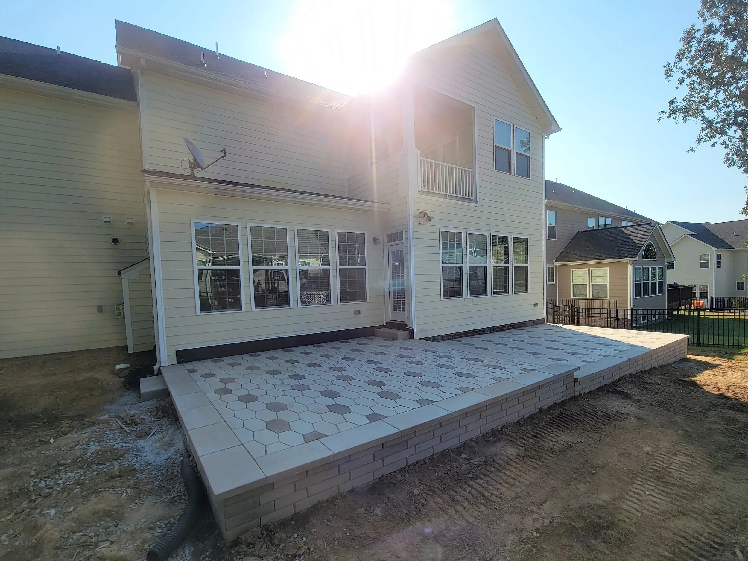 Backyard patio with a hexagon tile pattern and a house with beige siding, large windows, a balcony, and a satellite dish under a bright sun.