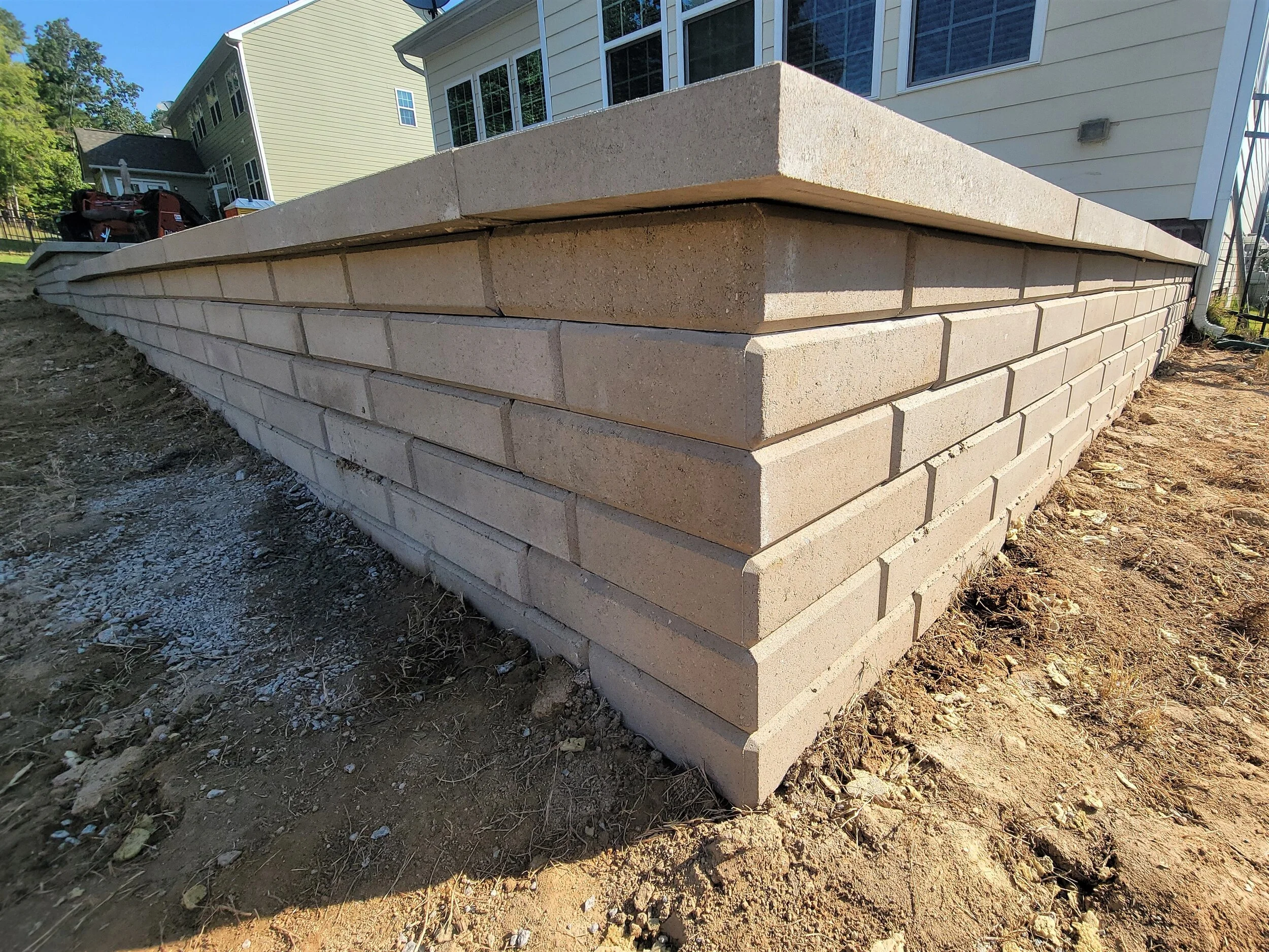 A newly constructed brick retaining wall with a concrete cap, adjacent to a house with white siding and multiple windows, on a dirt ground.