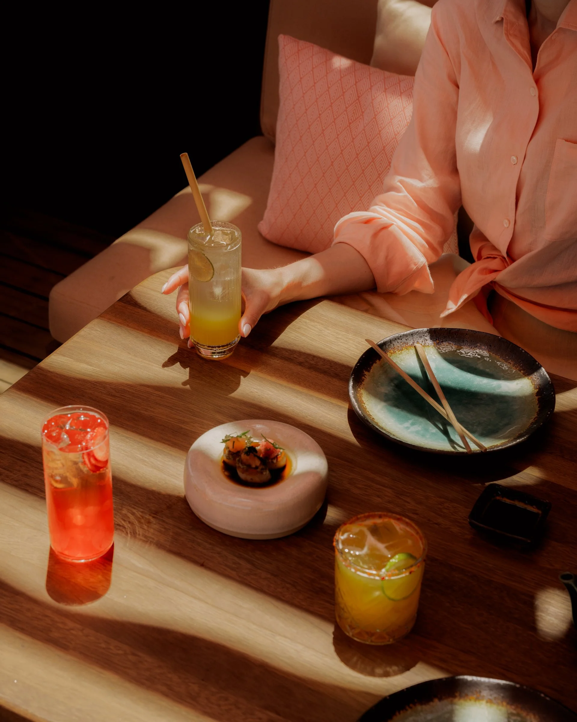 A woman in a pink shirt sitting at a wooden table with colorful drinks and a sushi dish, with shadows of window blinds on the table.