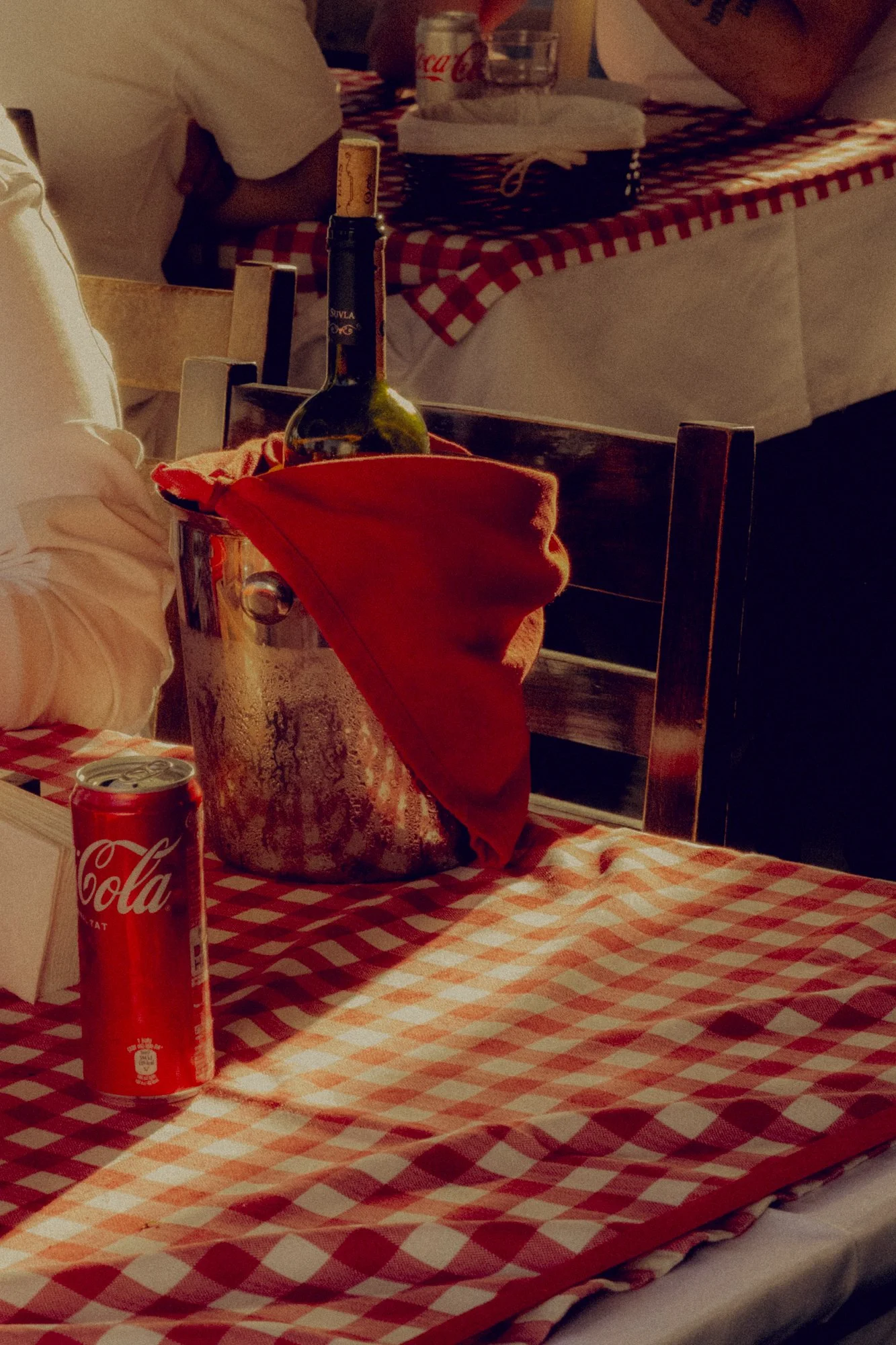 A table with a red and white checkered tablecloth, featuring a wine bottle in a metal ice bucket with a red cloth, a can of Coca-Cola, and some napkins. In the background, people are seated at another table with similar tablecloths.