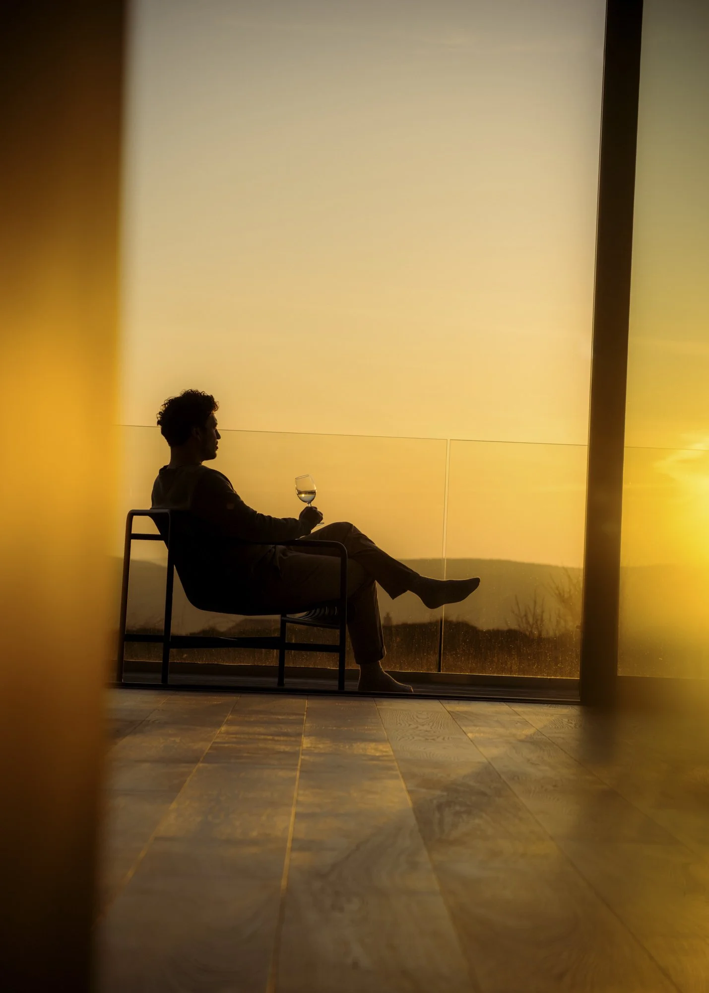 Person sitting on a chair, silhouetted against a sunset window, holding a glass of wine.