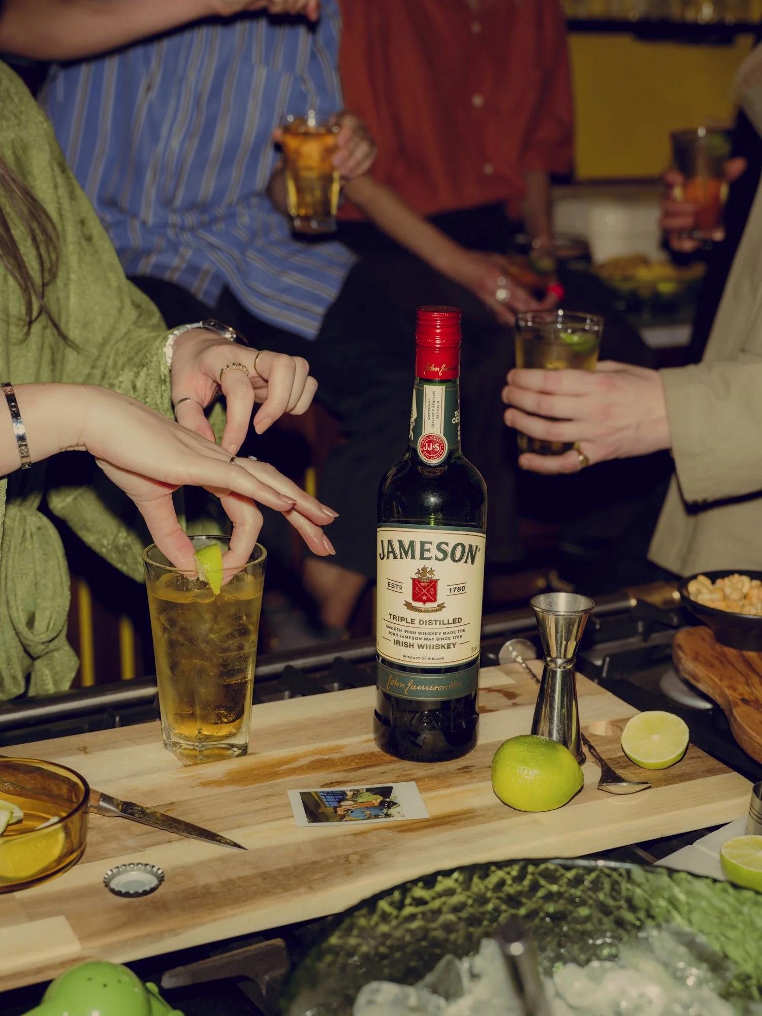 People holding drinks around a bar with a bottle of Jameson Irish whiskey, a lime, a muddler, and a photo on a cutting board.