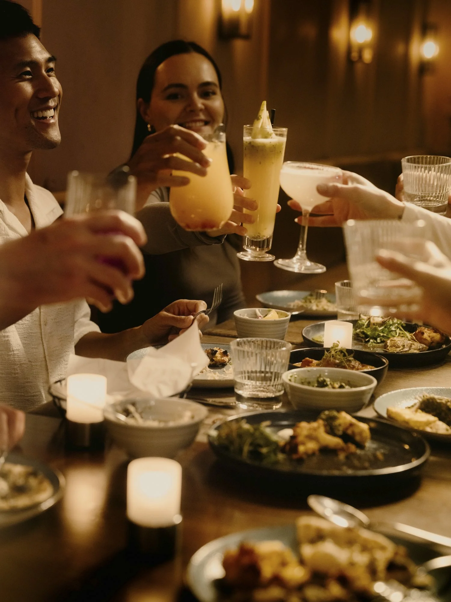People clinking drinks at a dinner party, surrounded by plates of food and candles on a wooden table in a warmly lit setting.