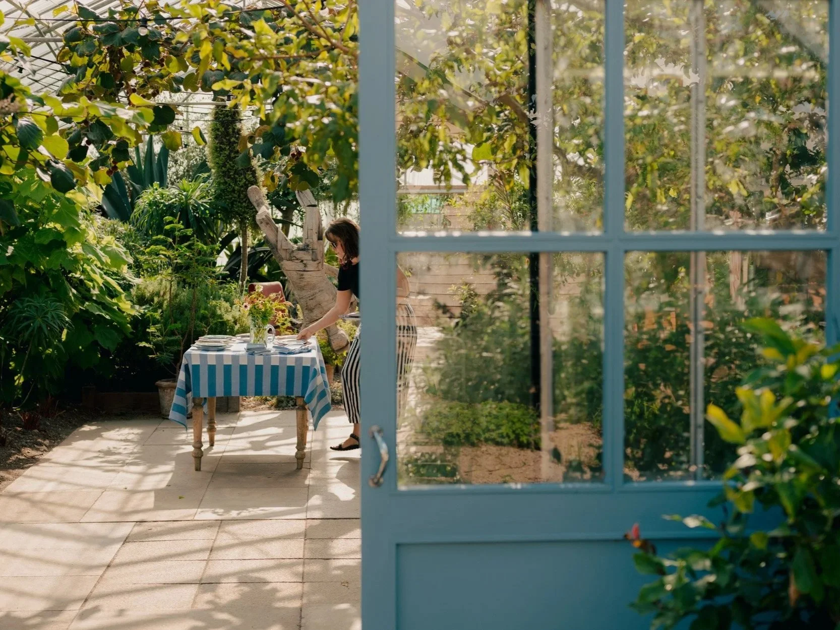 A woman setting a table with a blue and white striped tablecloth in a lush garden, seen through a partly open blue door.