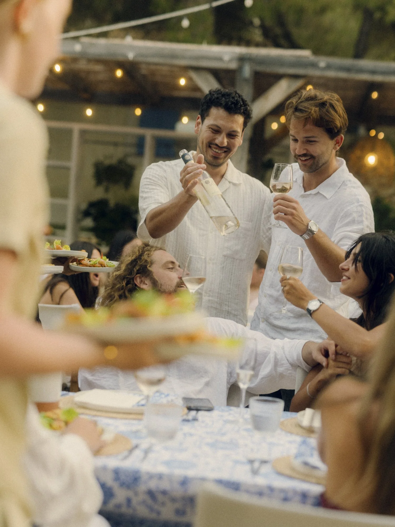 People celebrating at an outdoor party, with two men pouring wine and others enjoying food and drinks, under string lights.