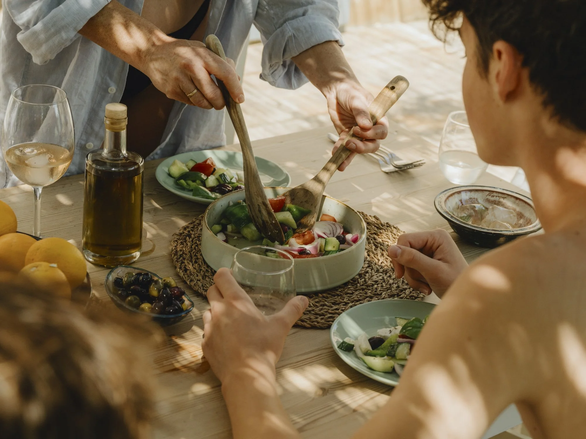 People enjoying a Mediterranean-style meal with fresh salad, wine, and olives at a wooden table.