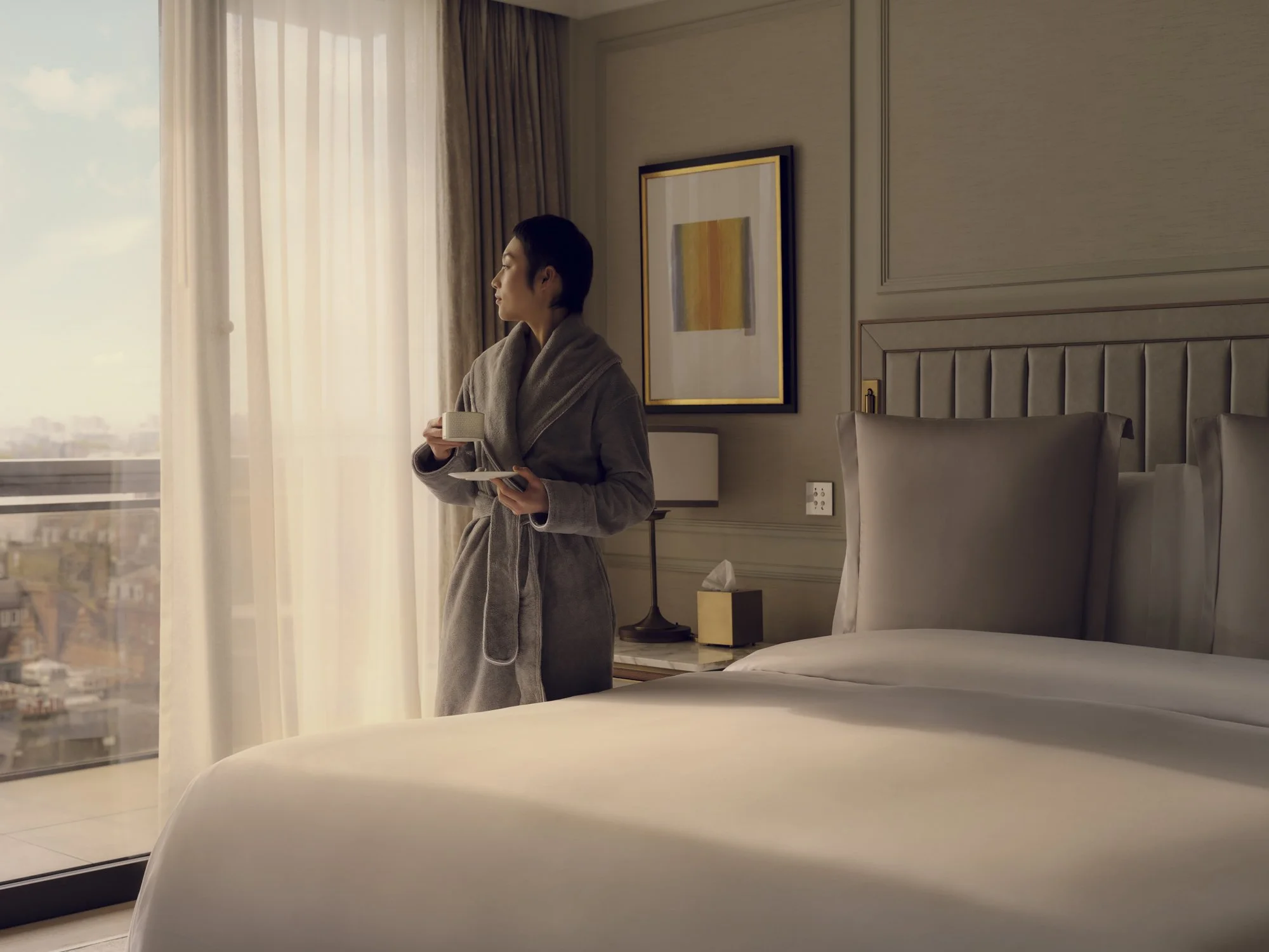 A woman with short dark hair wearing a gray bathrobe and holding a cup and a phone, standing by a large window with sheer curtains in a hotel room, looking outside.