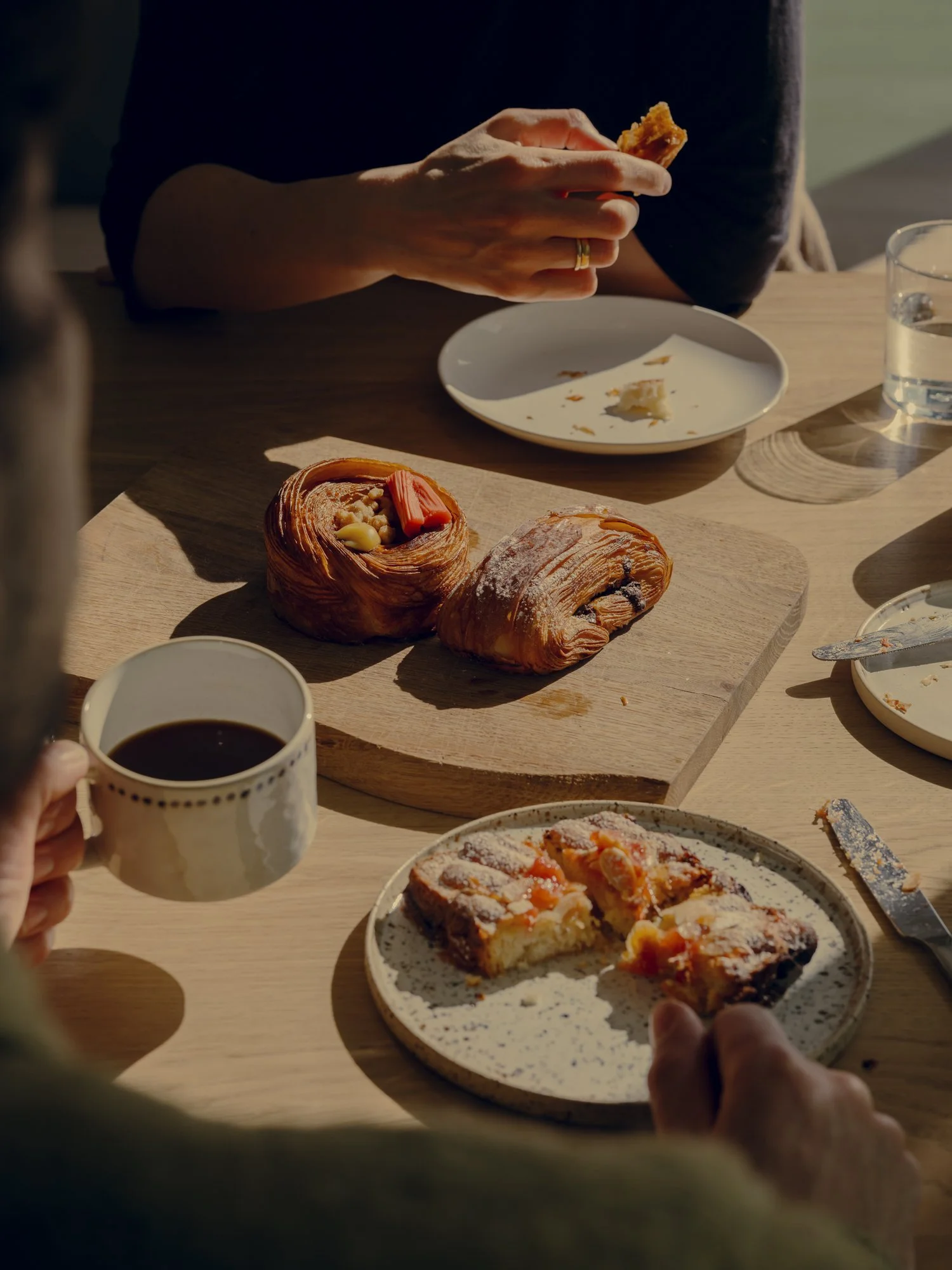 A table with pastries, a cup of coffee, and a glass of water, with people sharing a meal in natural lighting.