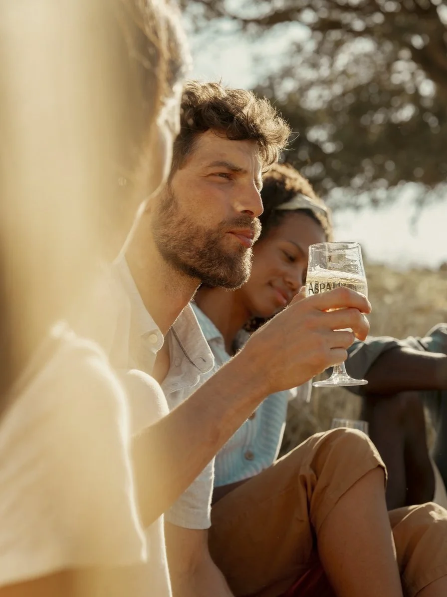 A group of young friends sitting outdoors, with one man holding a glass of beer or cider, enjoying a sunny day.