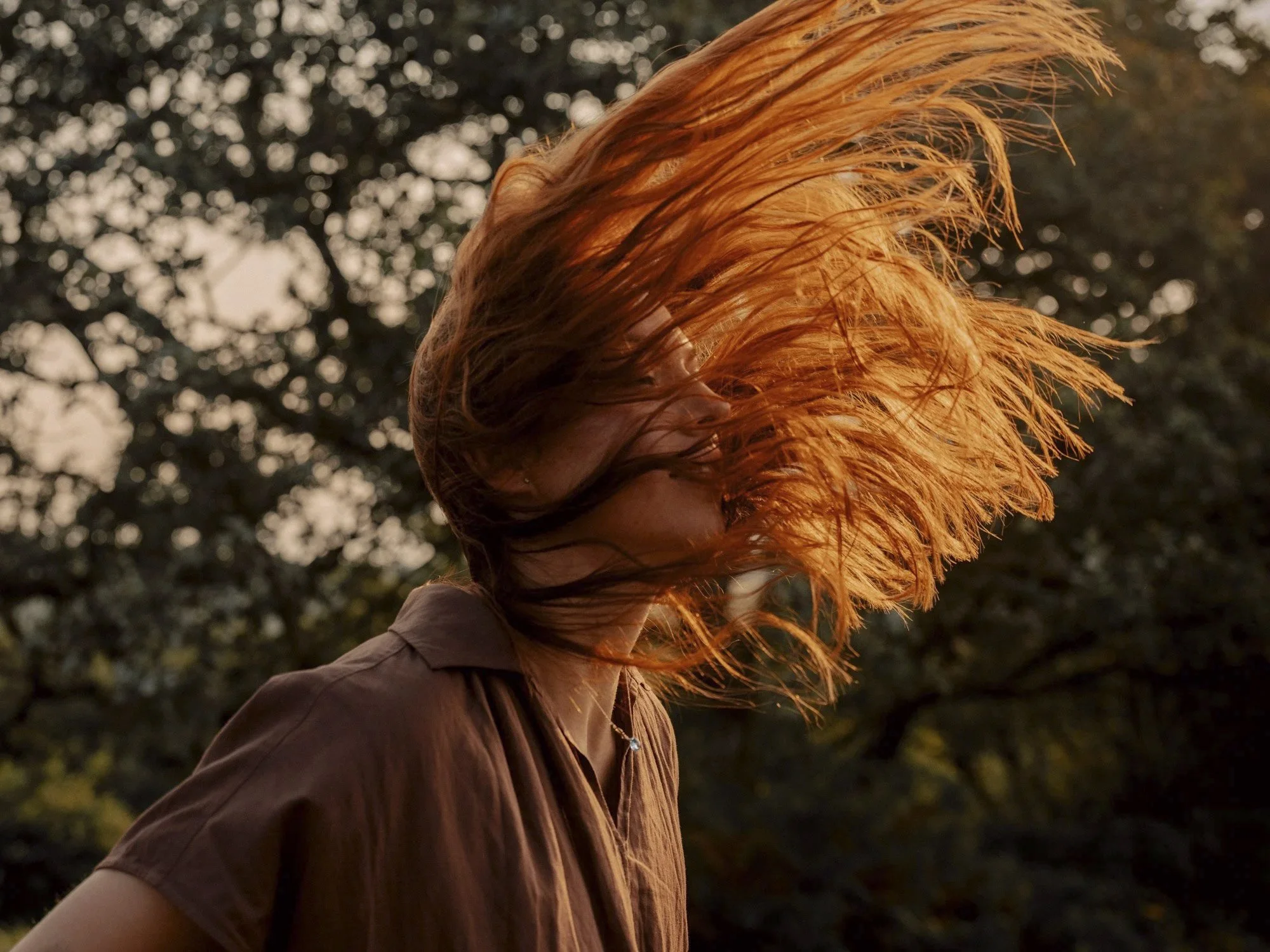 A person with long red hair flipping their hair in a park during sunset, with trees in the background.