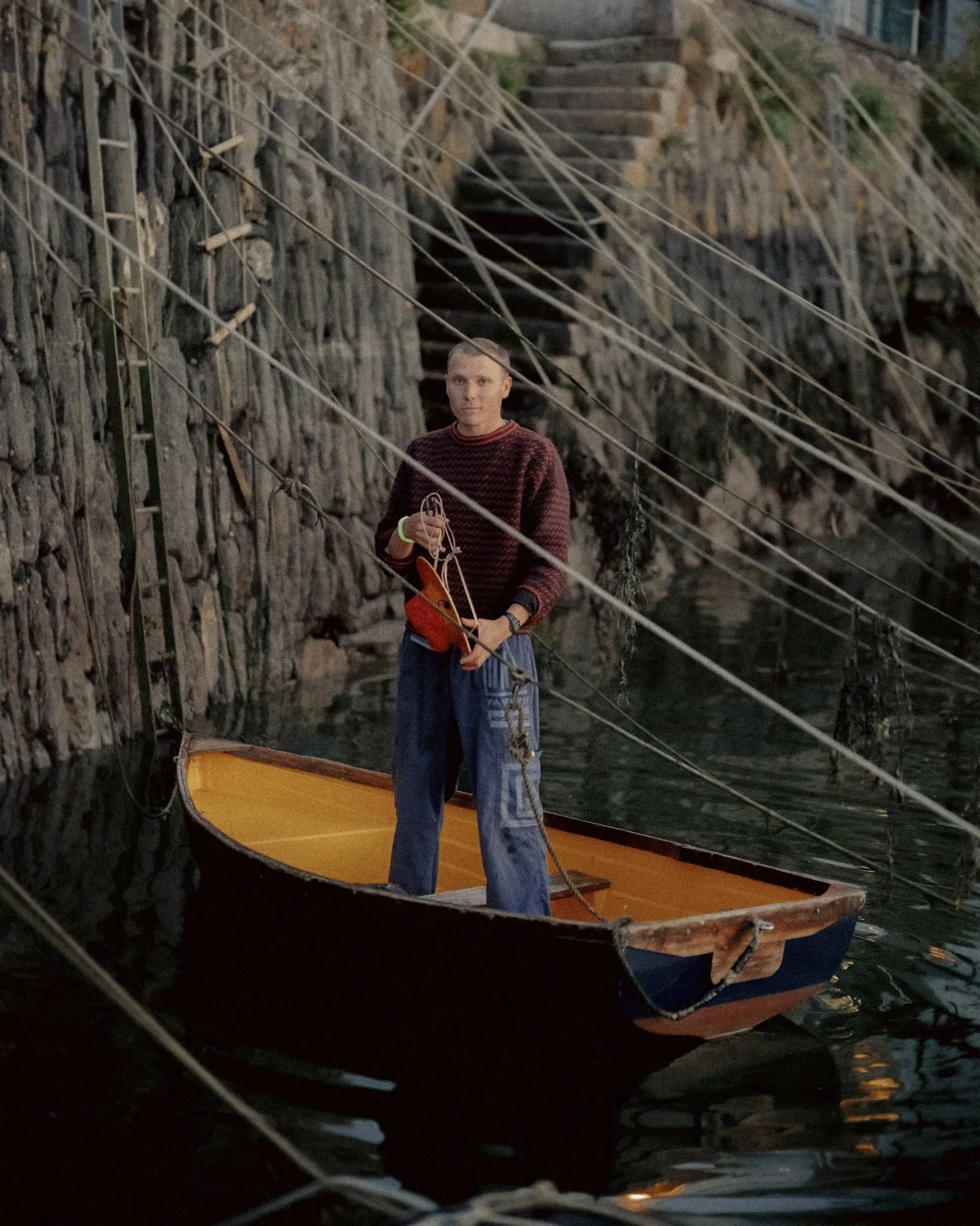 A person standing in a small wooden boat on a narrow waterway, with rocky walls and stairs in the background.