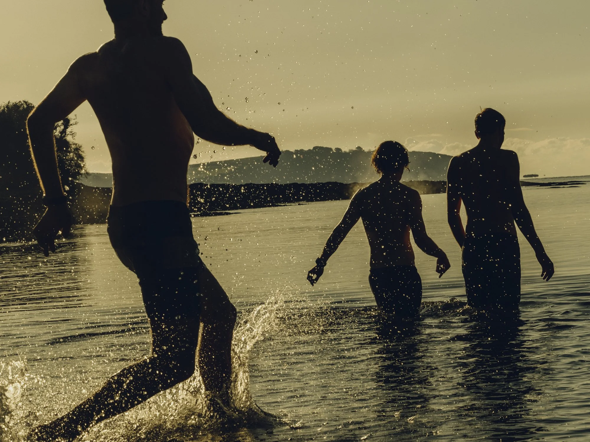 Silhouettes of three people walking and running in the water at sunset or sunrise.