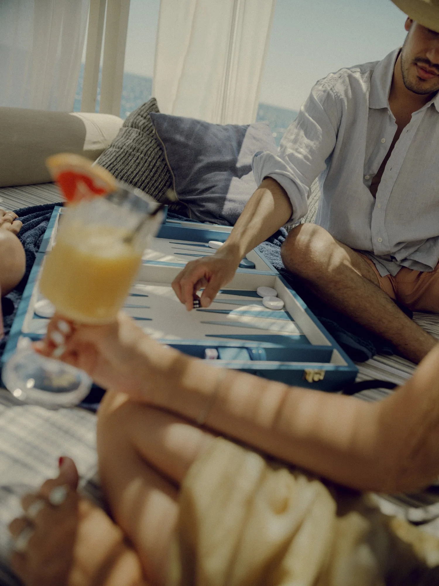 People playing backgammon on a deck near the ocean, with drinks and snacks, enjoying a sunny day.