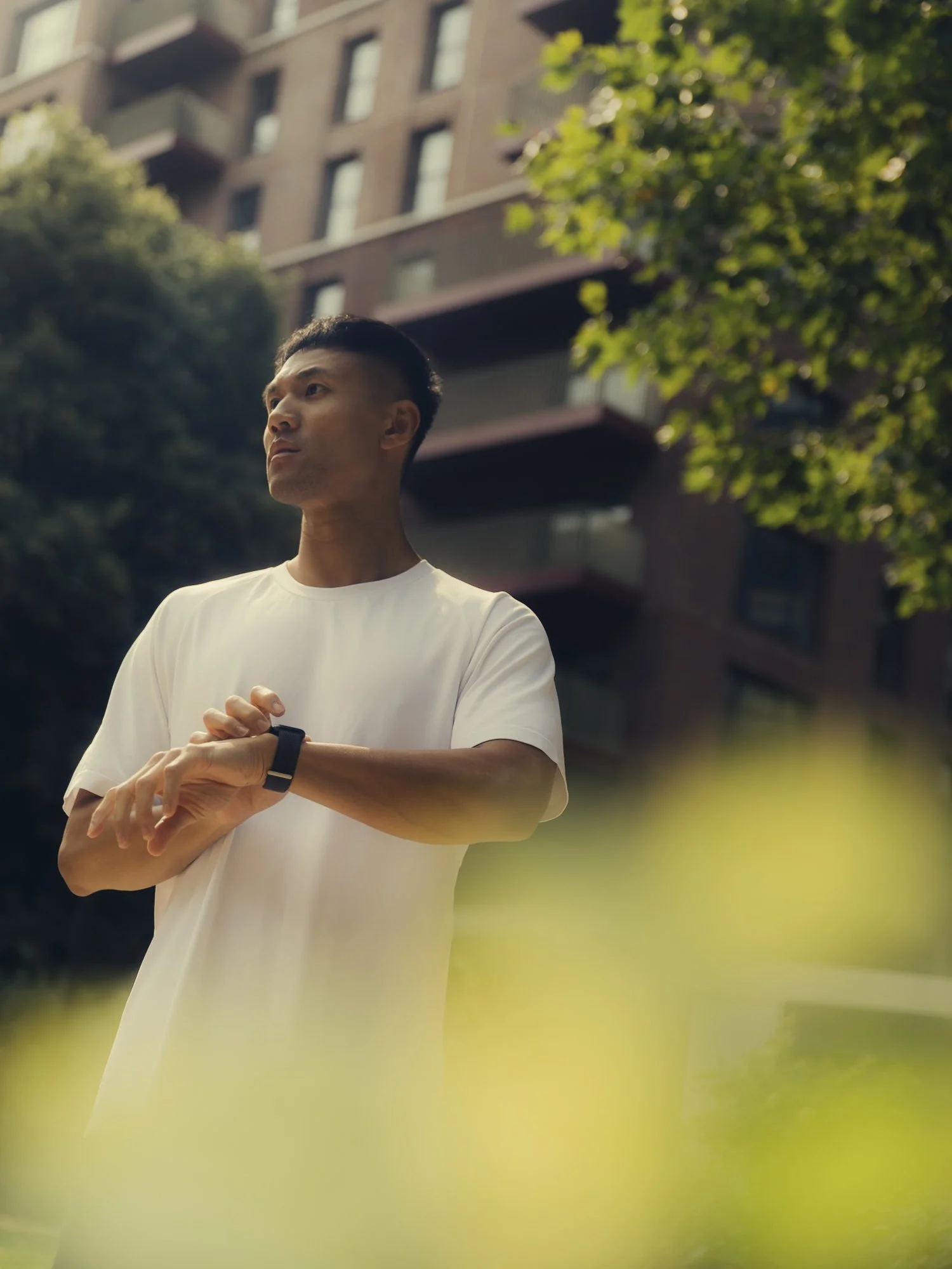 A man in a white T-shirt looking at his watch outdoors with trees and a multi-story building in the background.