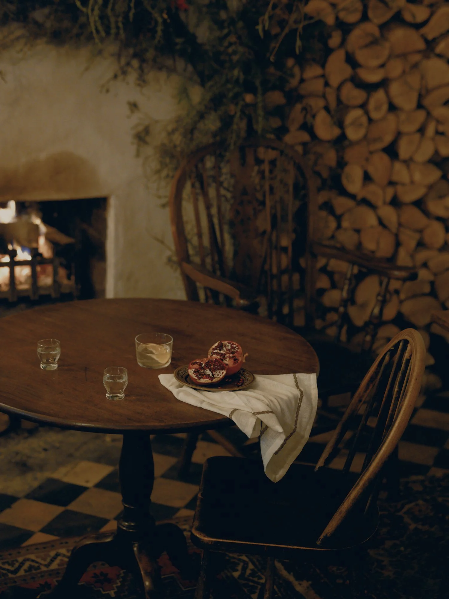 A cozy dining scene with a dark wooden table set near a fire, with a white napkin, a plate of pomegranate seeds, and three glasses of wine or juice. Wooden chairs surround the table, and a wall of stacked firewood is visible in the background.
