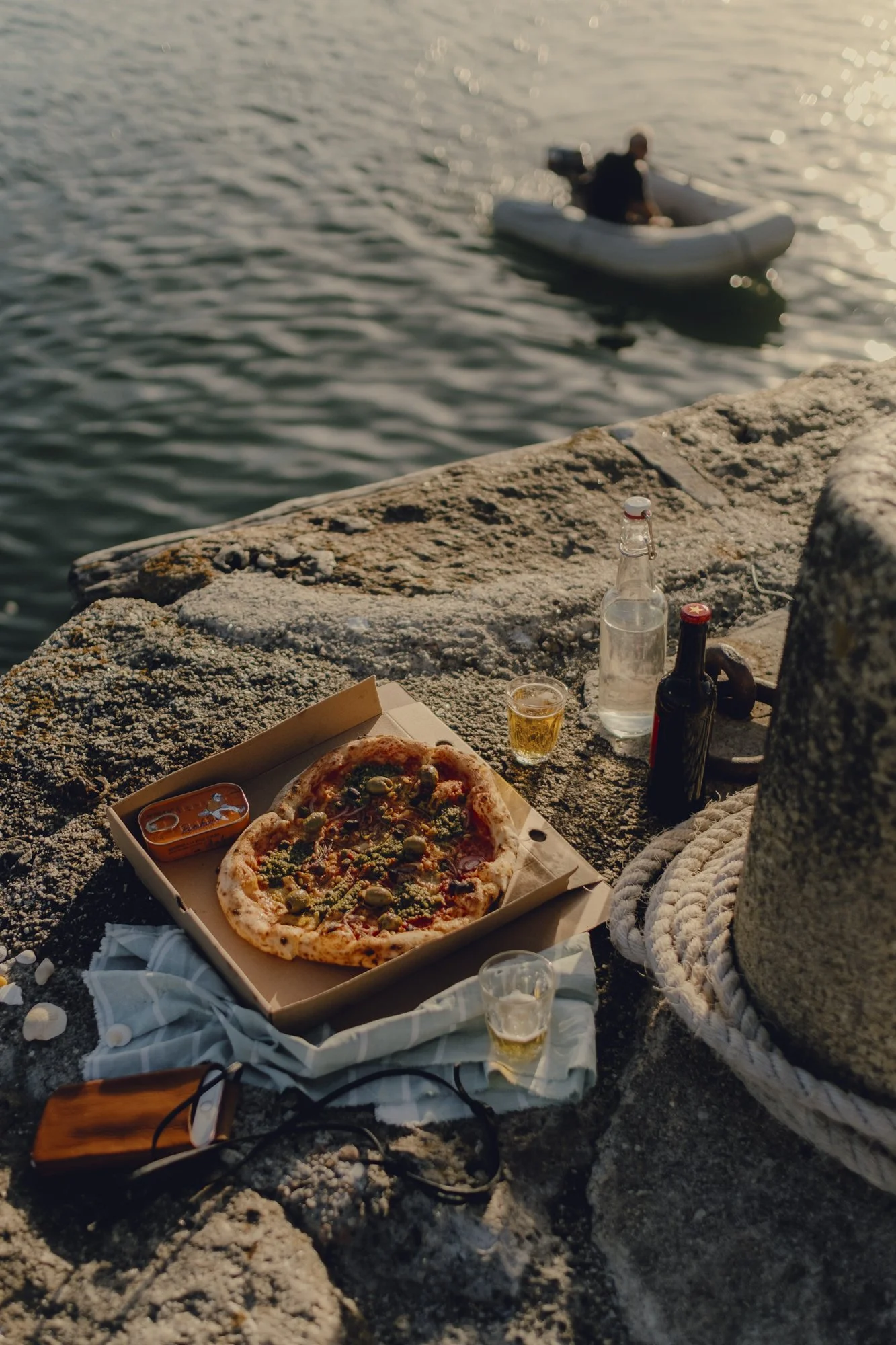 Pizza box with pizza, two beers, water bottle, and small glasses, with a person in a boat on the water in the background.