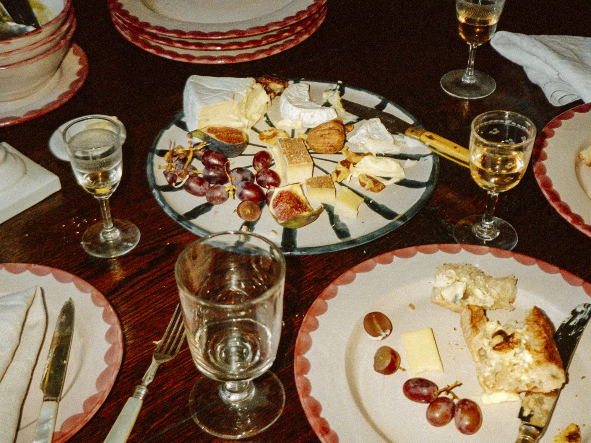 Cheese platter with grapes, figs, and nuts on a table with glasses of white wine and plates with leftover cheese and grapes.