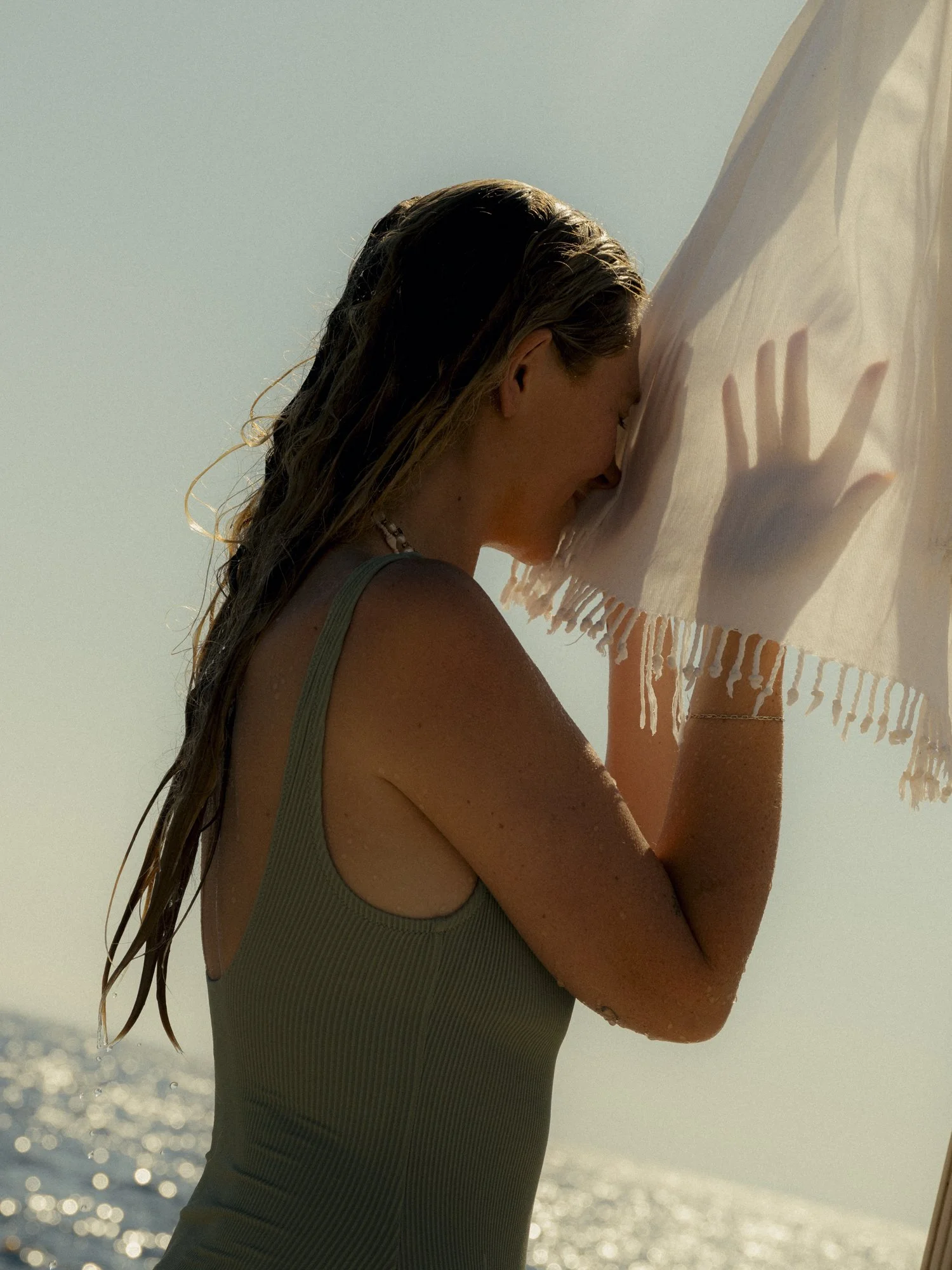 Woman with wet hair smiling and holding a white towel near the ocean on a sunny day.